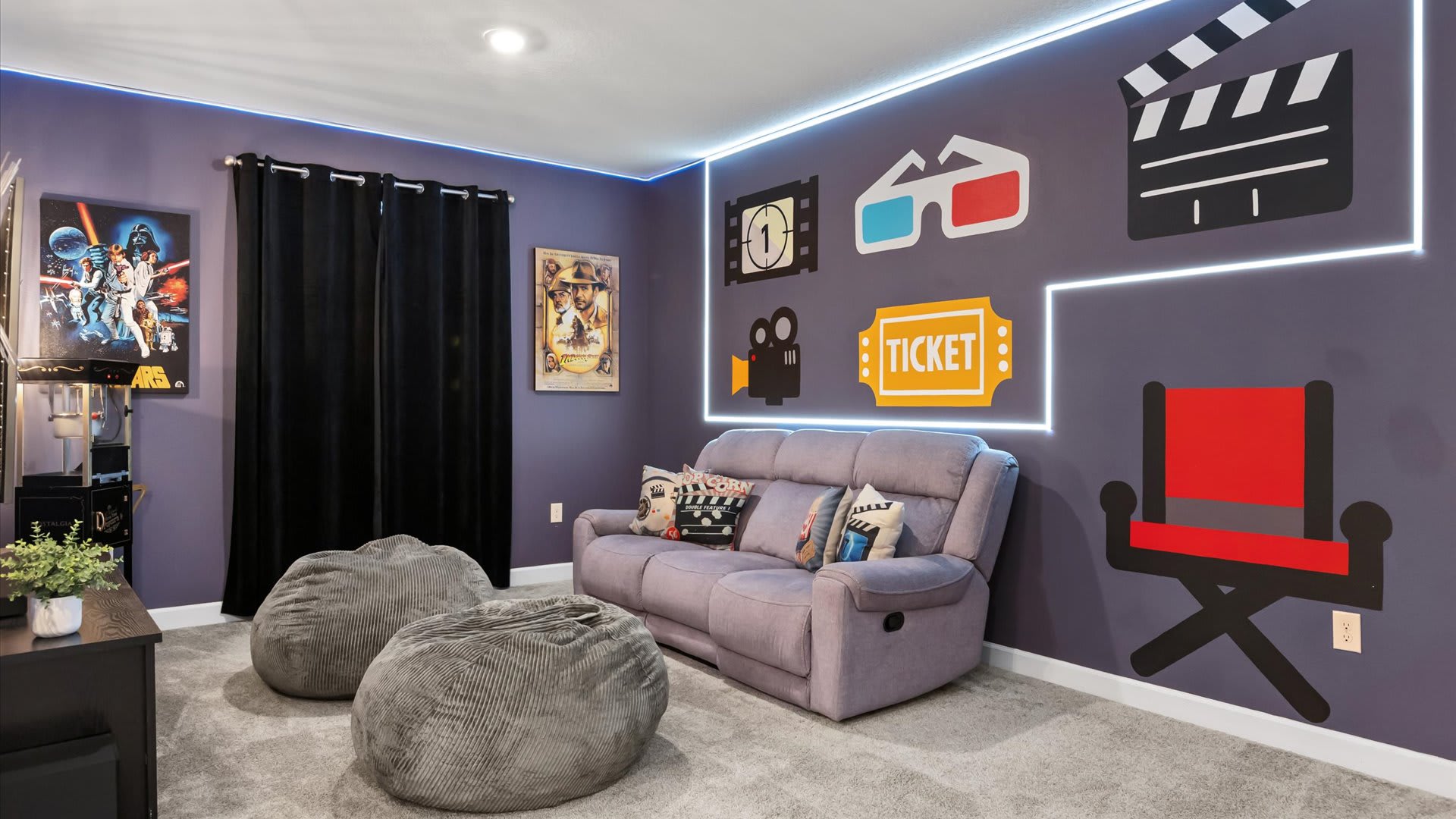 A lively media room with purple walls decorated with movie-themed decals and white LED strip lighting, offering a grey reclining sofa and two grey bean bag chairs for comfortable viewing. Black curtains frame a window, and a vintage Indiana Jones poster adds to the cinematic ambiance.