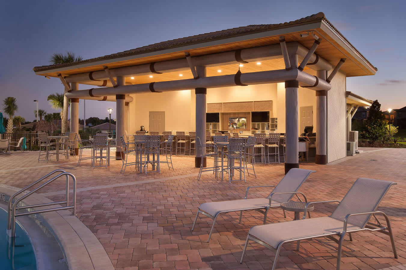Outdoor covered bar and lounge area next to a swimming pool, illuminated at dusk. The space features high-top tables, chairs, and lounge chairs on a brick patio, with a bar counter in the background.