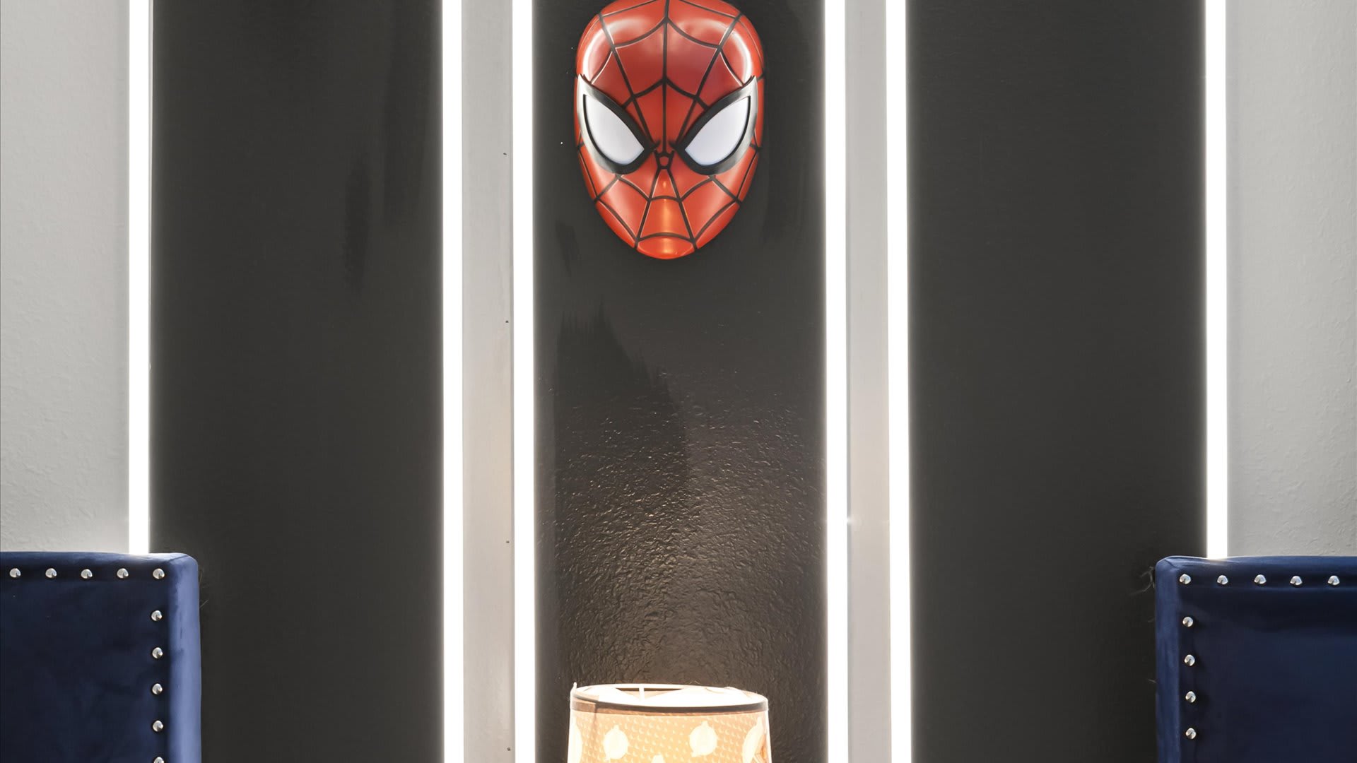 Close-up of a dramatic black accent wall with vertical white LED strips, featuring a prominent Spiderman mask decoration. A small table lamp is visible below.