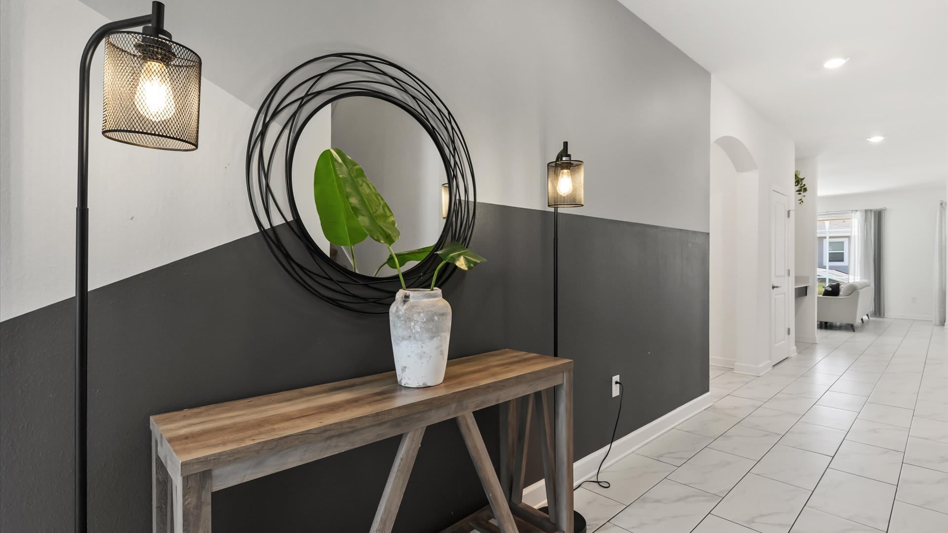 A stylish hallway features a two-tone wall with a dark gray lower half and white upper half, adorned with a large circular black mirror and a wooden console table. Two tall black floor lamps with caged shades illuminate the space, leading into other areas of the home.