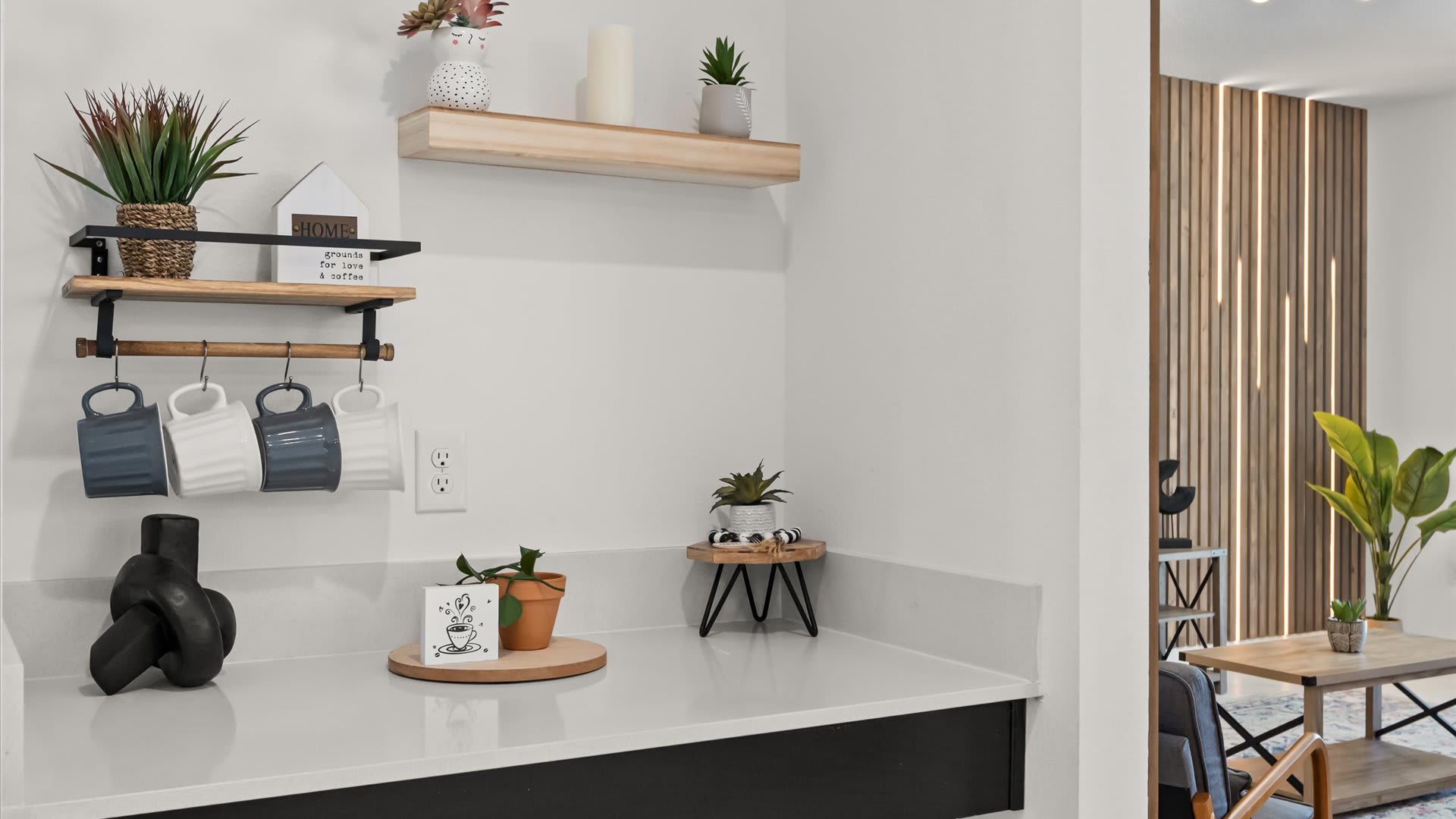 A dedicated coffee bar nook in the kitchen features a wooden mug rack with four mugs, a floating shelf with decorative plants and candles, and a white countertop. The space is adorned with a "HOME" sign and small potted plants, offering a cozy beverage station.