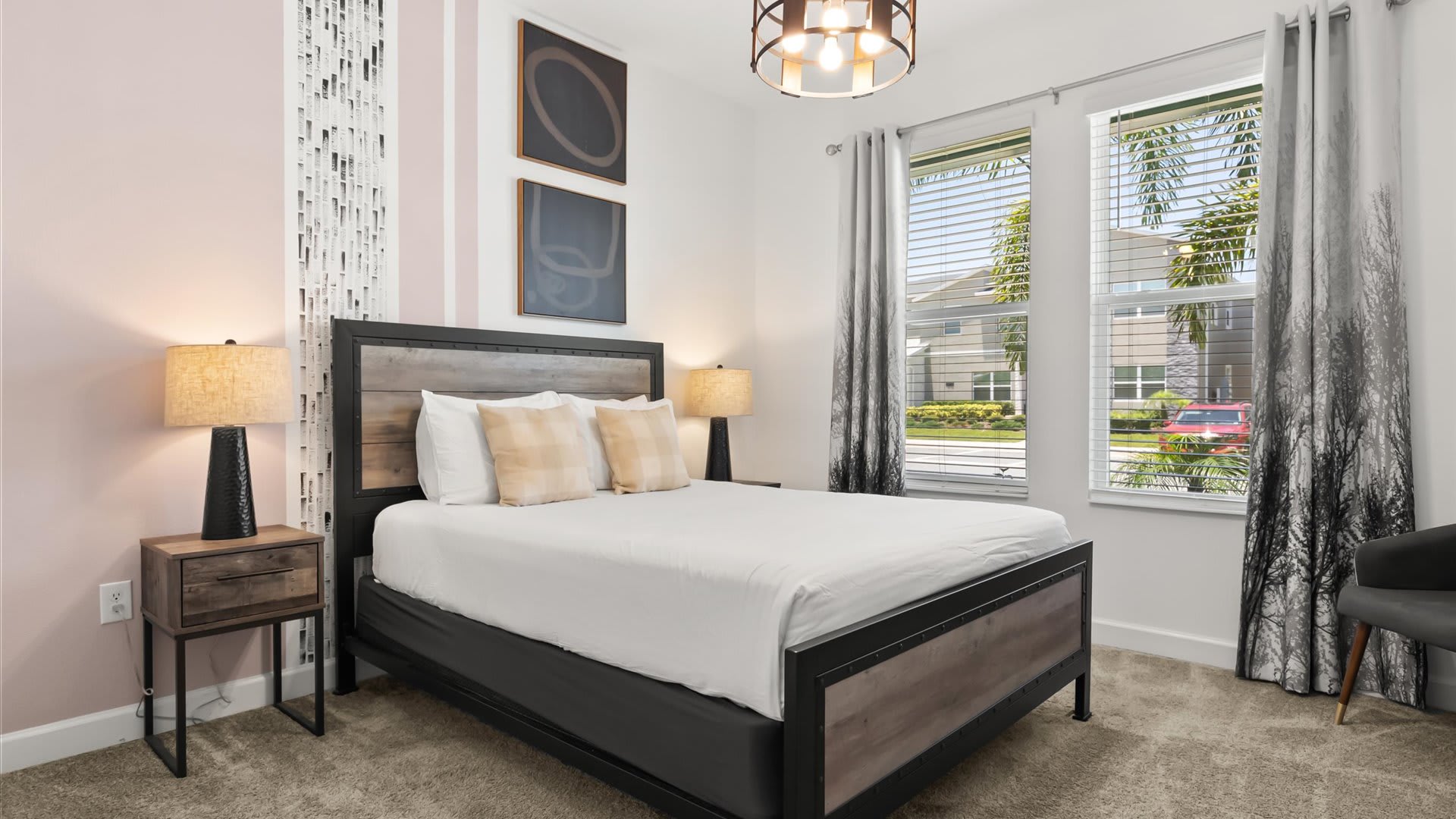 Cozy bedroom featuring a rustic wood and metal bed frame, two matching nightstands with table lamps, and large windows with blinds and patterned curtains. A light pink accent wall adds a touch of color.