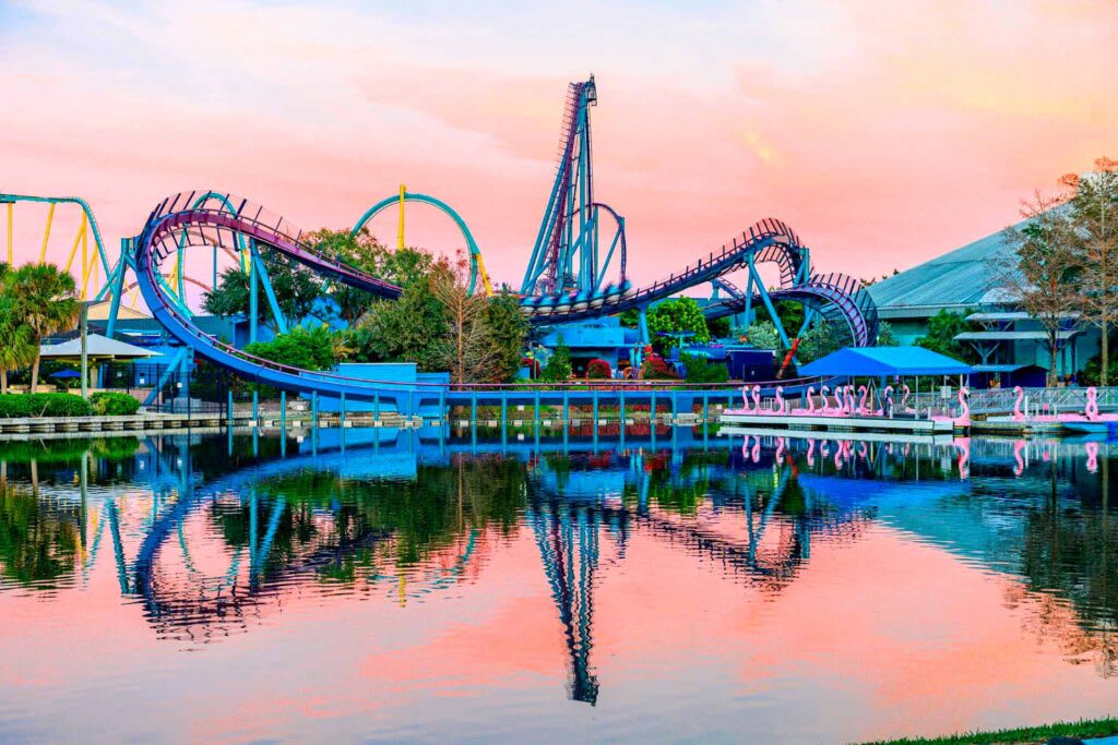 A vibrant sunset over a theme park lake, reflecting a large blue roller coaster with multiple loops and drops, surrounded by lush greenery and a pink-orange sky.