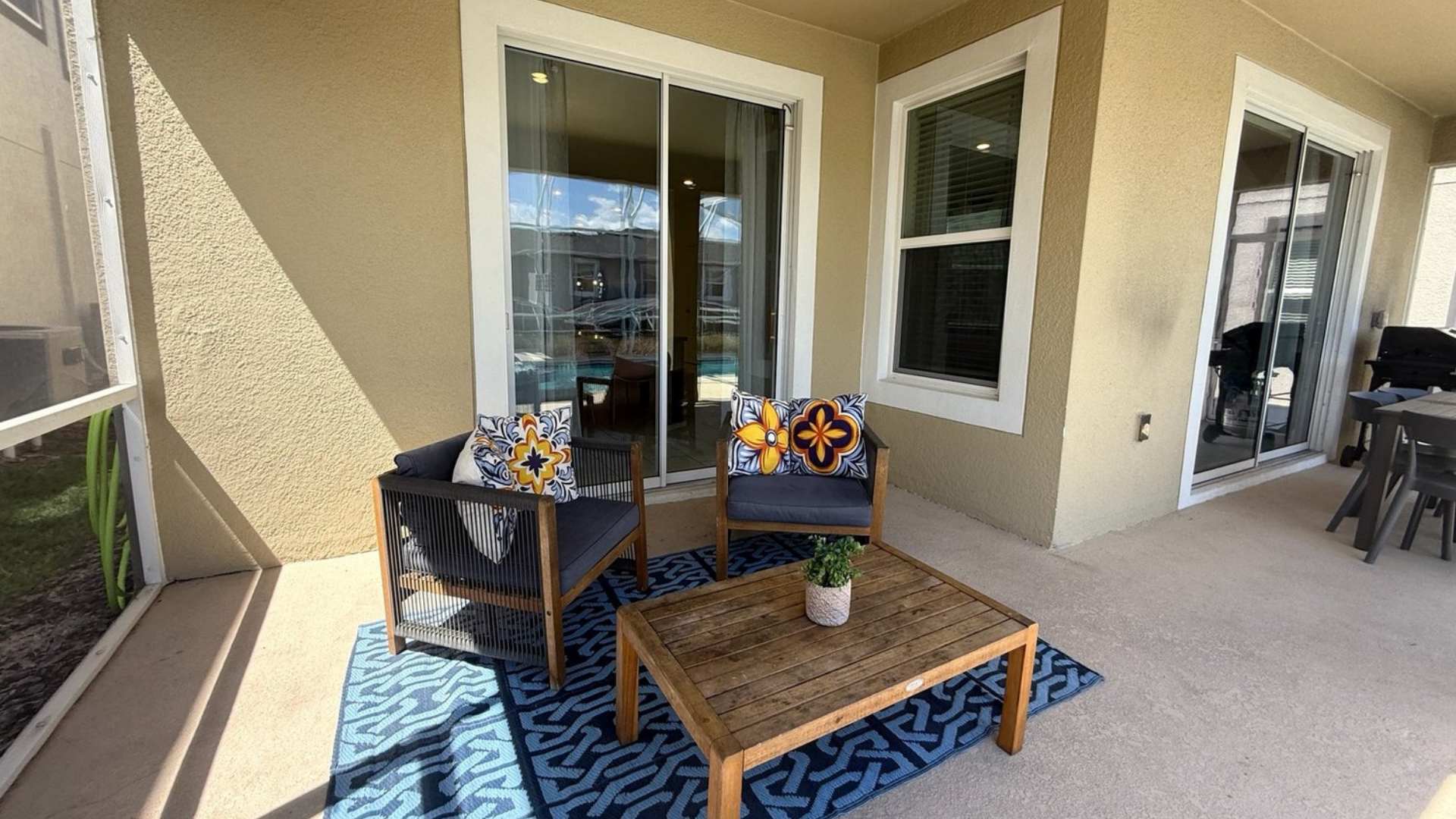 Covered outdoor patio area with two comfortable armchairs and a wooden coffee table on a blue patterned rug, offering a view of the screened pool area through sliding glass doors.