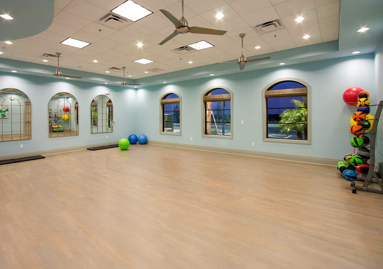 A bright and spacious fitness studio with light blue walls, light wood flooring, and multiple arched windows. It features large mirrors, ceiling fans, recessed lighting, and a rack of colorful exercise balls.