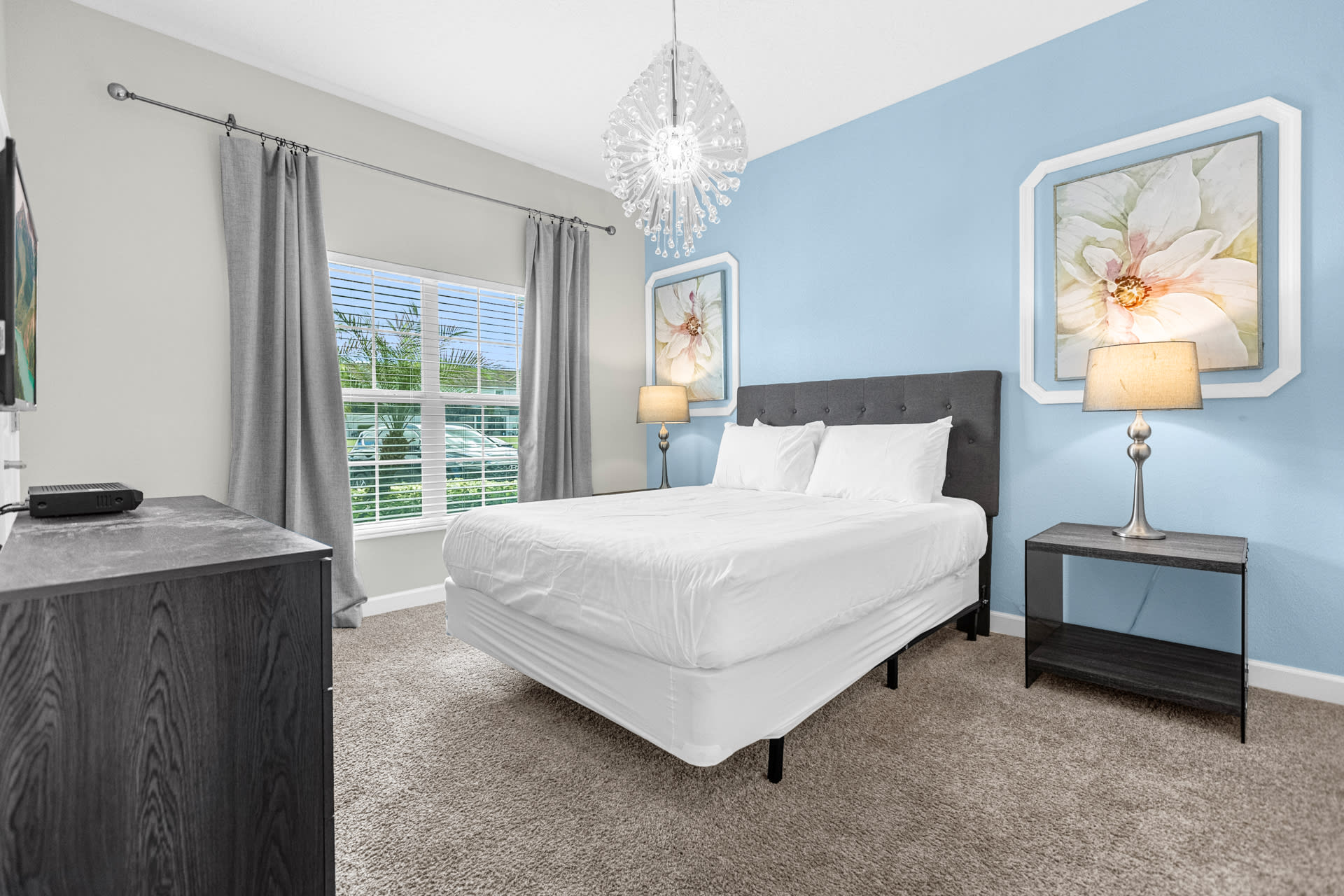 Cozy bedroom featuring a queen-sized bed with a dark gray headboard, a light blue accent wall with decorative floral art, and a large window with blinds and gray curtains, illuminated by a modern chandelier.