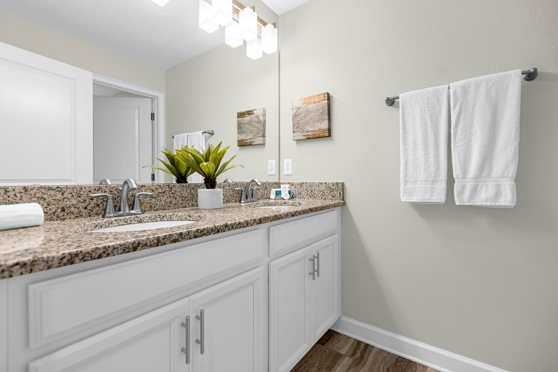Close-up of a double vanity in a bathroom, featuring granite countertops, white shaker cabinets, two sinks, and a large mirror, with fresh white towels and a green plant accent.