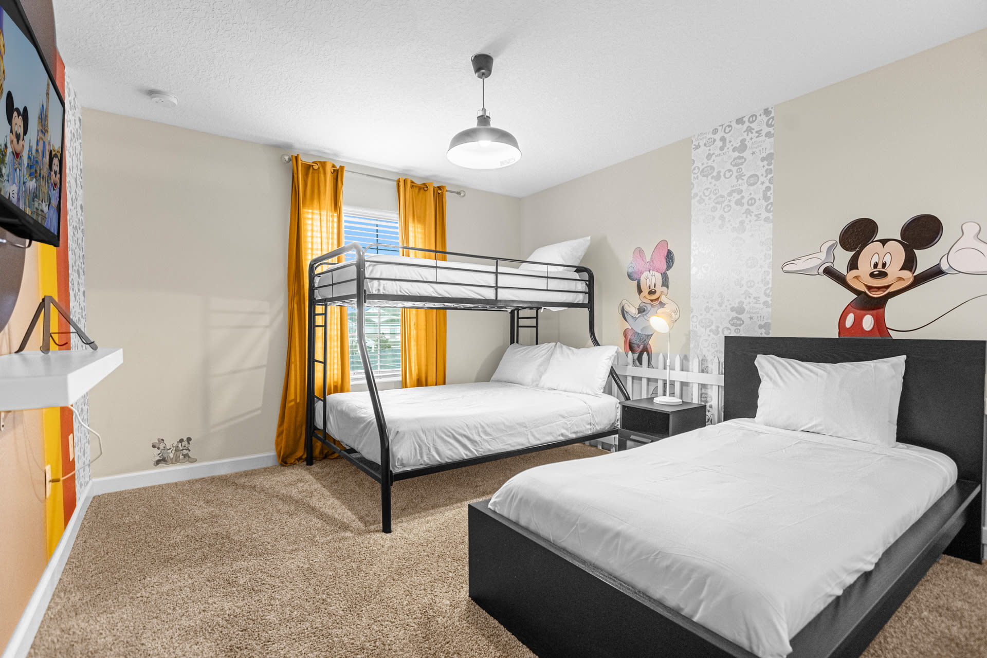 Bright kids' bedroom featuring a black metal bunk bed with white bedding, a matching single bed, and Mickey and Minnie Mouse wall decals. Yellow curtains frame a window, and a TV is mounted on a colorful stand, all set on a light brown carpet.