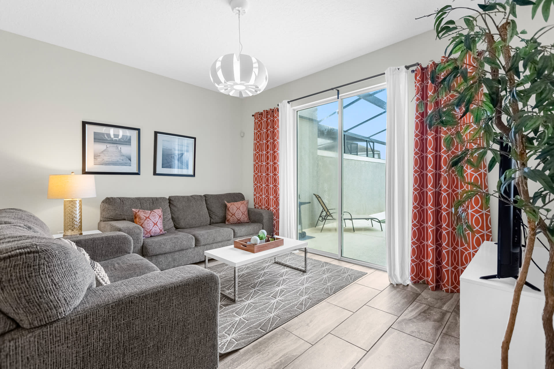 Bright living room with gray sofas, a white coffee table, and a large potted plant. Sliding glass doors with orange and white curtains provide access to the screened outdoor patio.