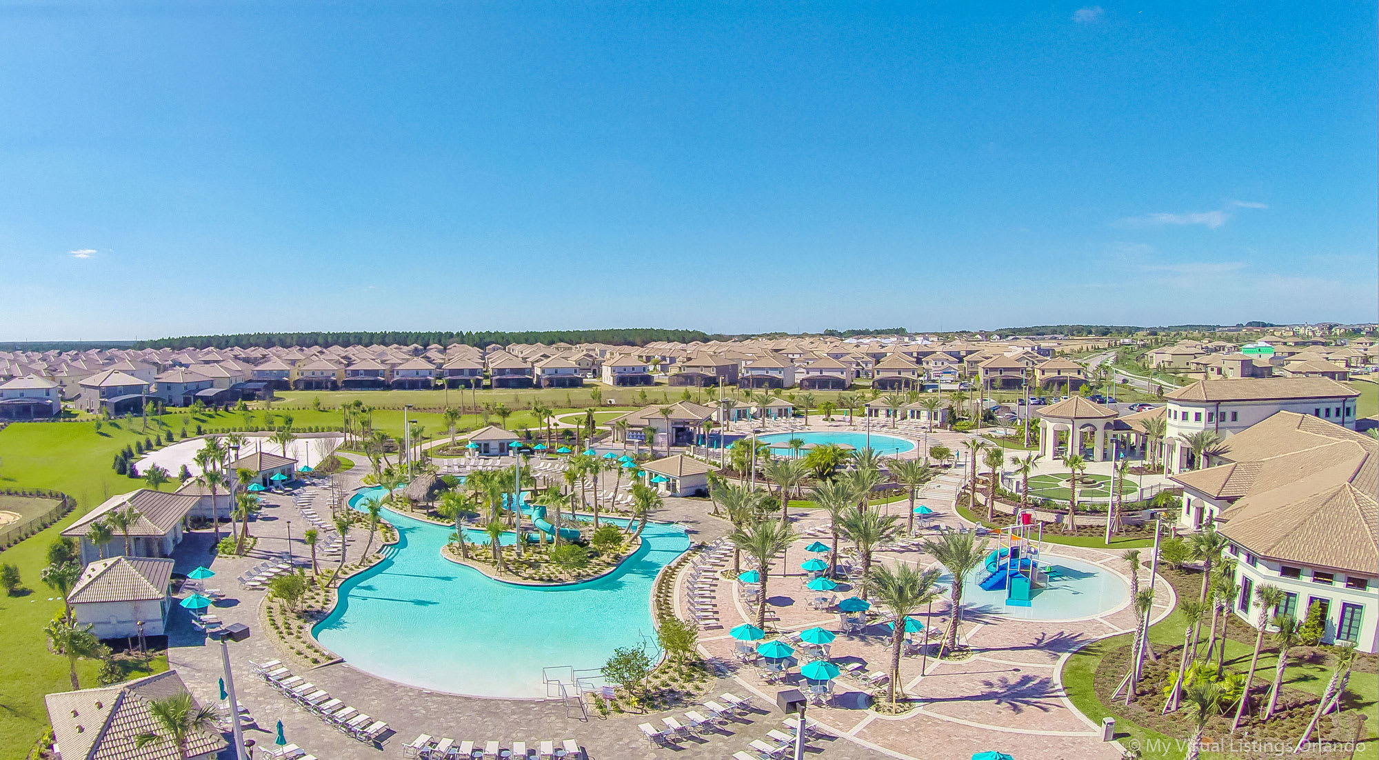 Expansive aerial view of a large resort community under a clear blue sky, showcasing multiple interconnected swimming pools, a central clubhouse, and numerous residential homes surrounded by green landscaping.