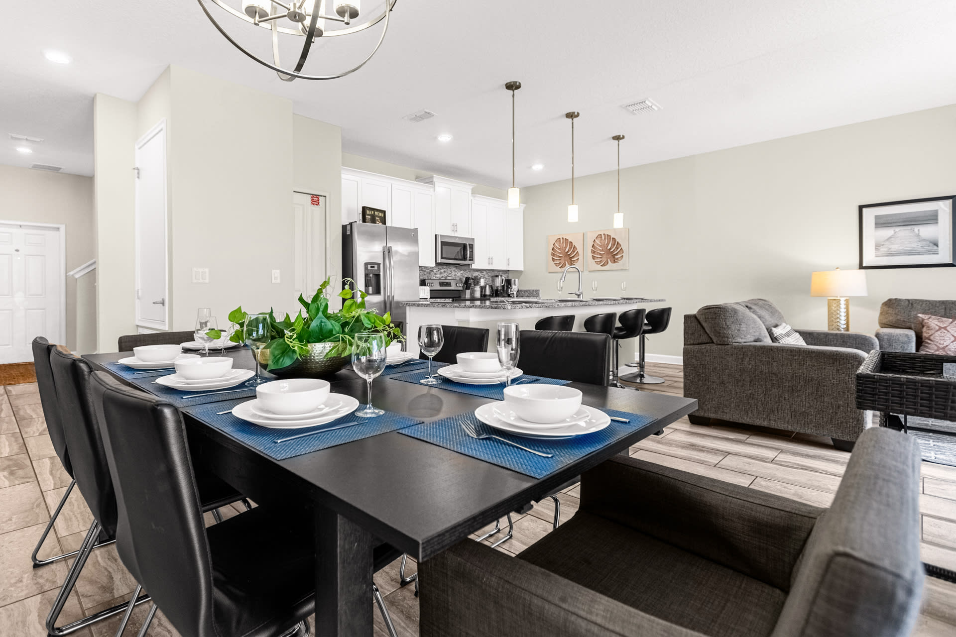 Modern dining area with a dark rectangular table set for eight, featuring blue placemats and white dinnerware, adjacent to a bright kitchen and living space with light walls and wood-look tile flooring.