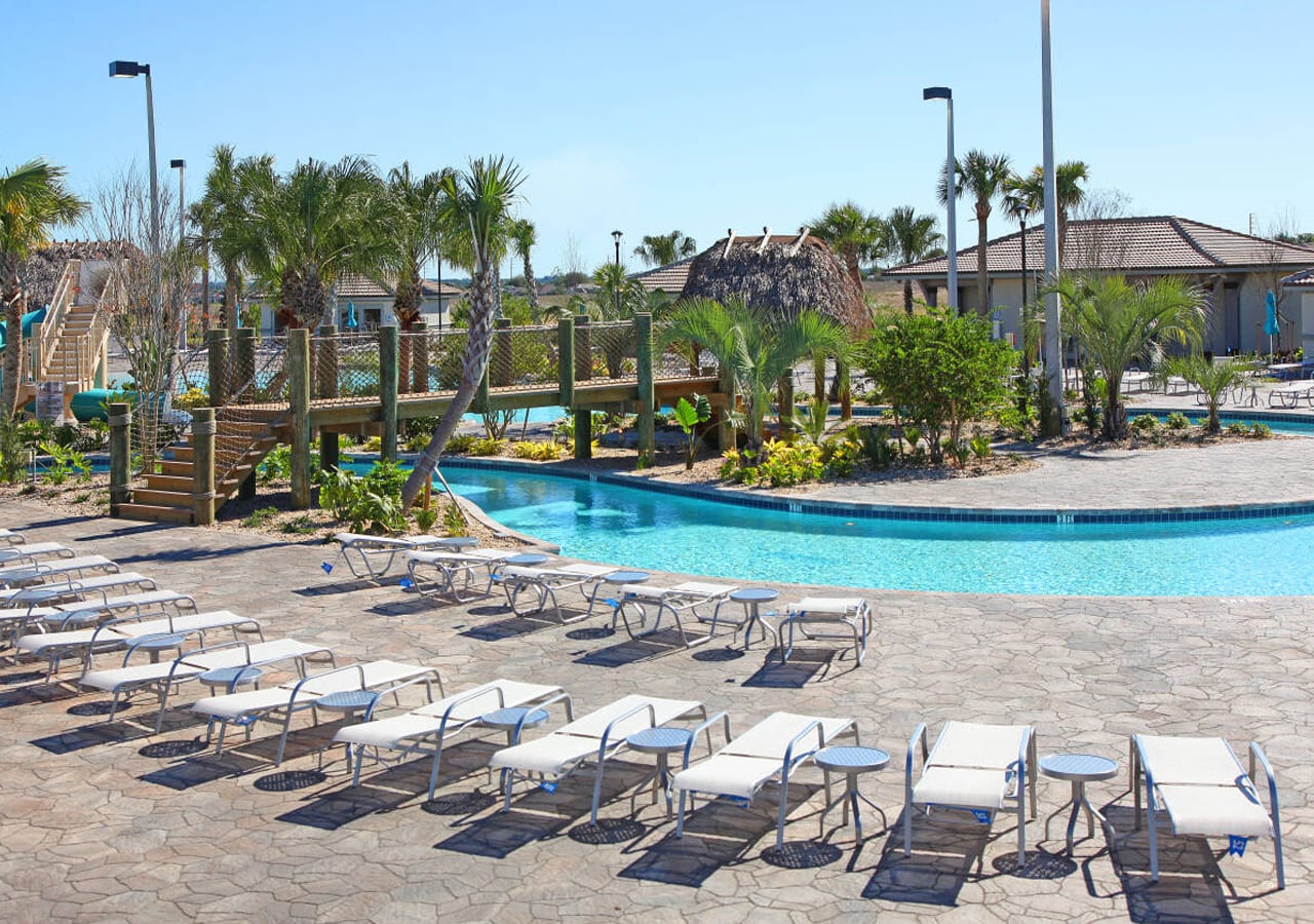 Resort pool area featuring a winding lazy river with a wooden bridge crossing over it. Numerous white lounge chairs are arranged on the stone-paved deck, surrounded by lush palm trees and tropical landscaping.