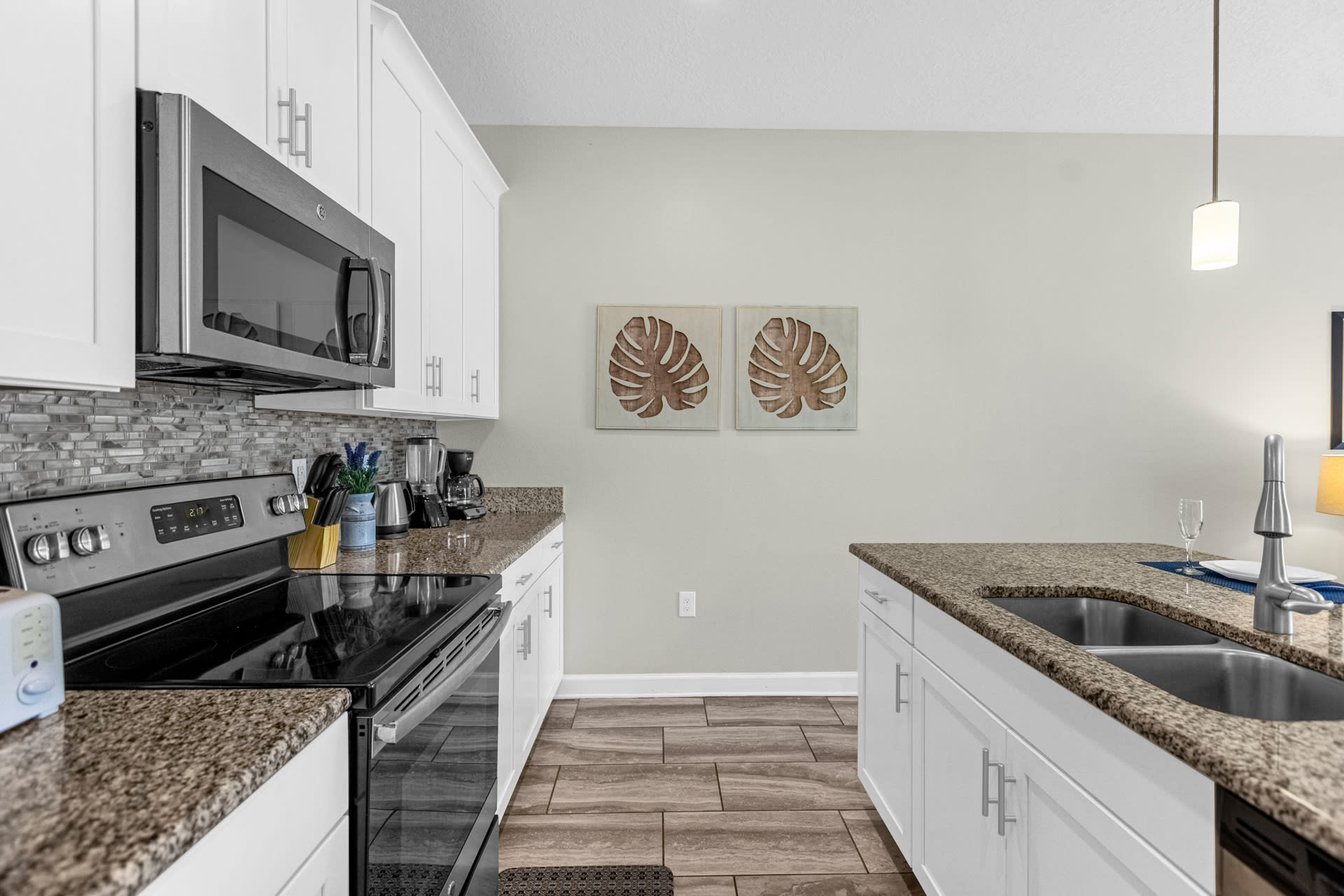 Modern kitchen with granite countertops, white shaker cabinets, a stainless steel electric range, microwave, and a gray tiled backsplash, featuring wood-look tile flooring and small appliances.