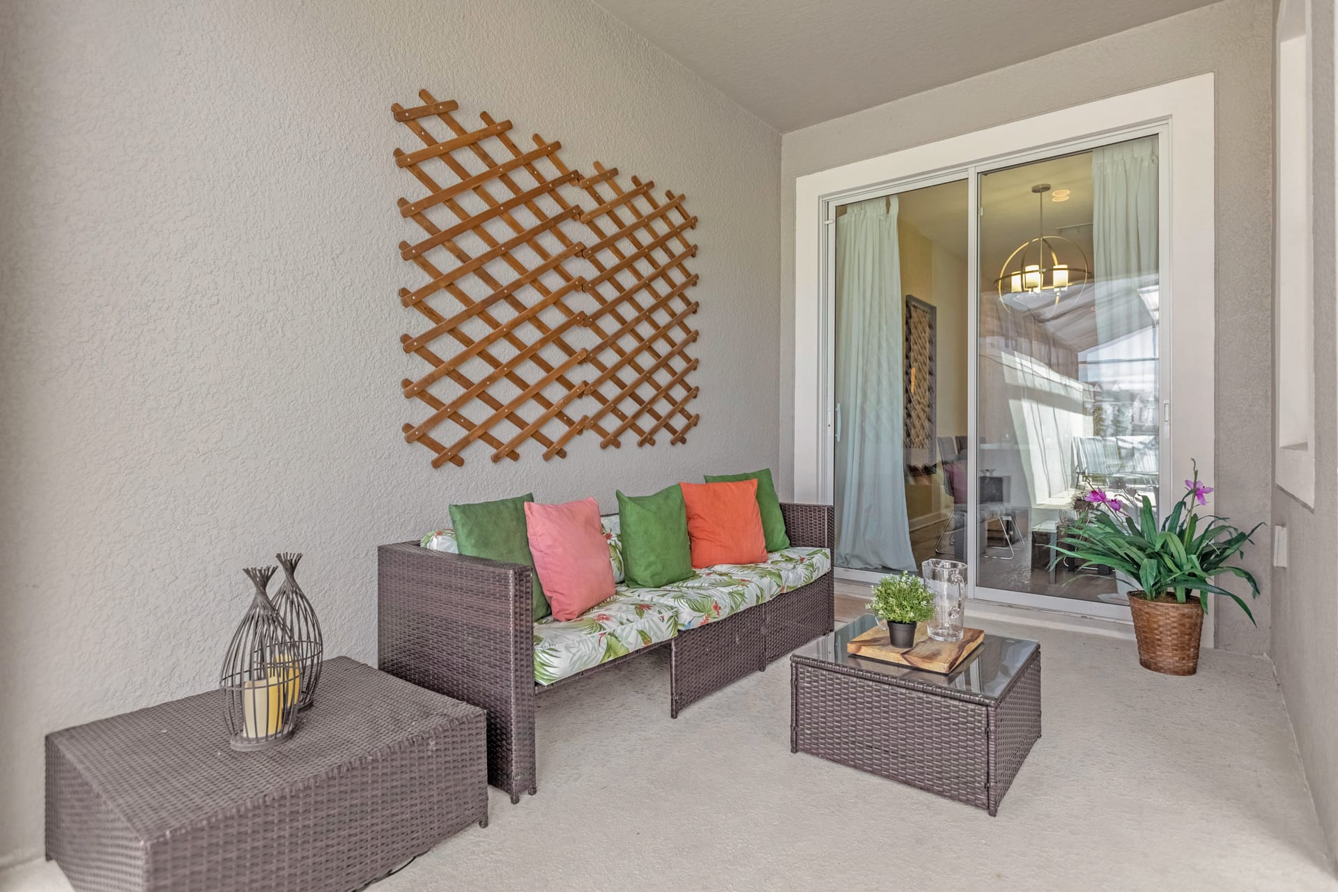 Covered outdoor patio with a wicker sofa adorned with colorful pillows and a matching coffee table. A decorative wooden trellis is mounted on the wall.