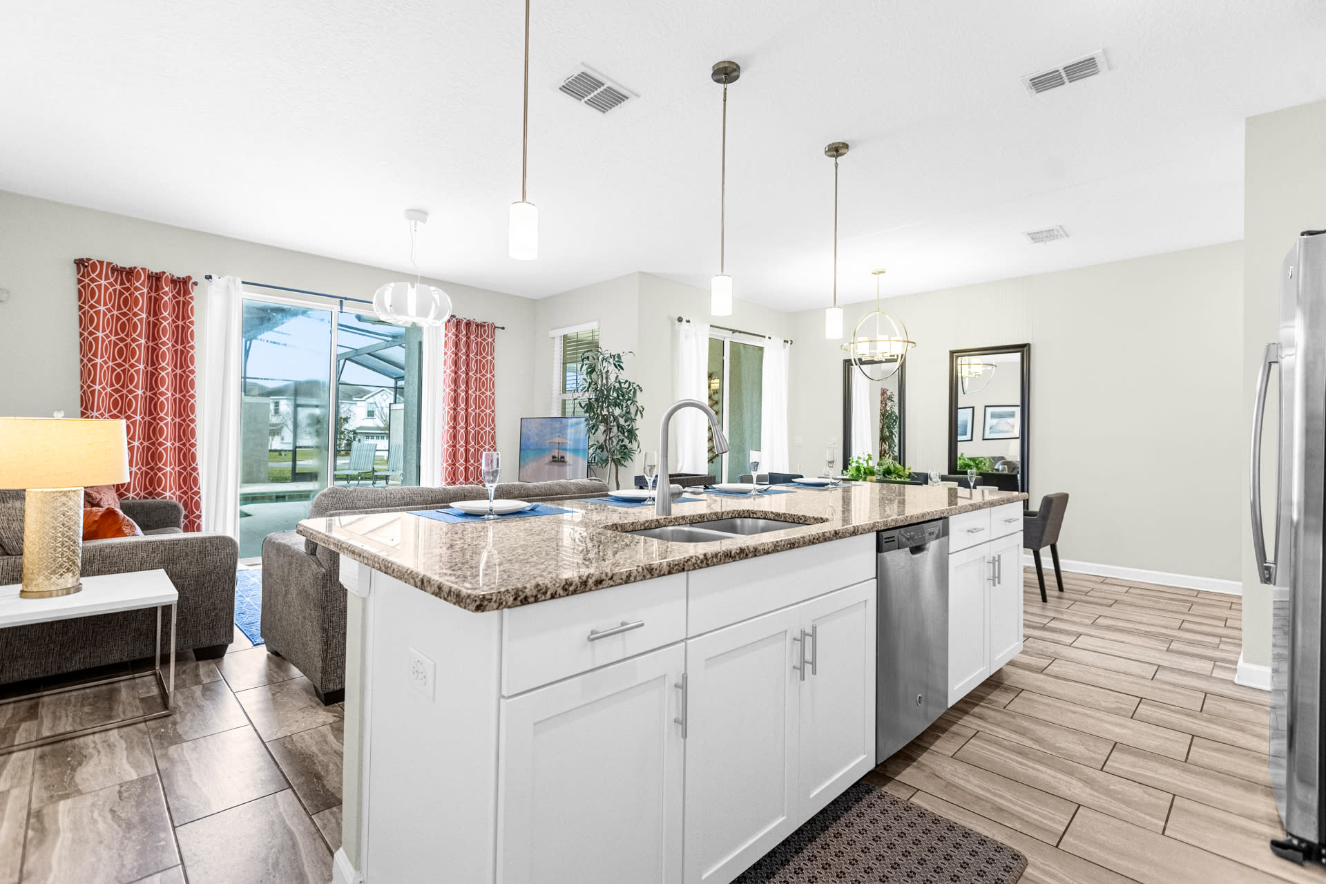 Expansive open-concept living space featuring a large kitchen island with granite countertops and a dishwasher, a comfortable living room with a gray sofa, and a dining area, all with access to an outdoor patio.