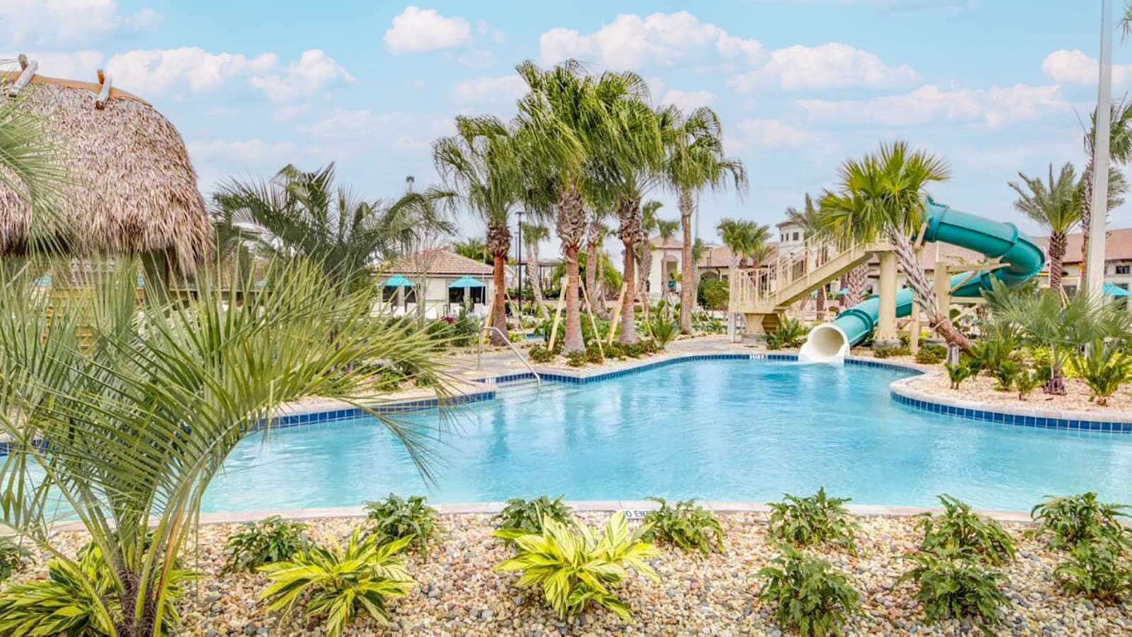 Tropical resort pool area with a large green water slide winding into the water. The pool is surrounded by lush palm trees, vibrant green plants, and decorative rocks under a bright, partly cloudy sky.