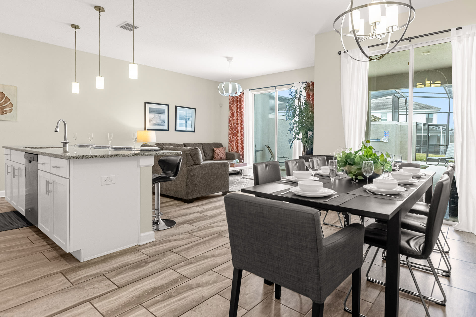 Open-concept dining area with a dark table and chairs, leading to a living room with a sofa and a kitchen island, all illuminated by natural light from a sliding glass door to an outdoor patio.