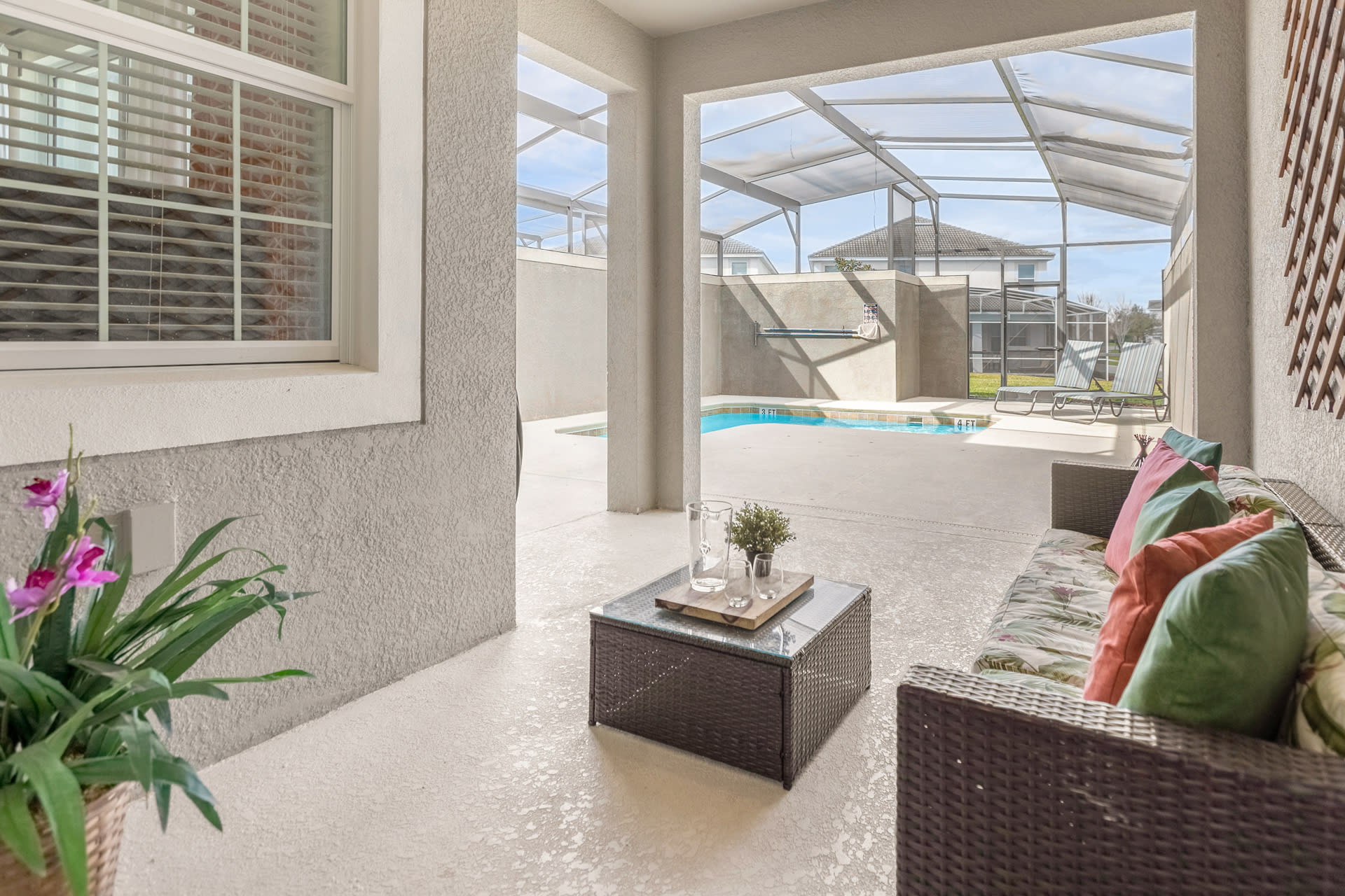 Covered outdoor patio area with wicker seating and a glass-top coffee table, offering a view of the screened private pool and surrounding concrete deck.
