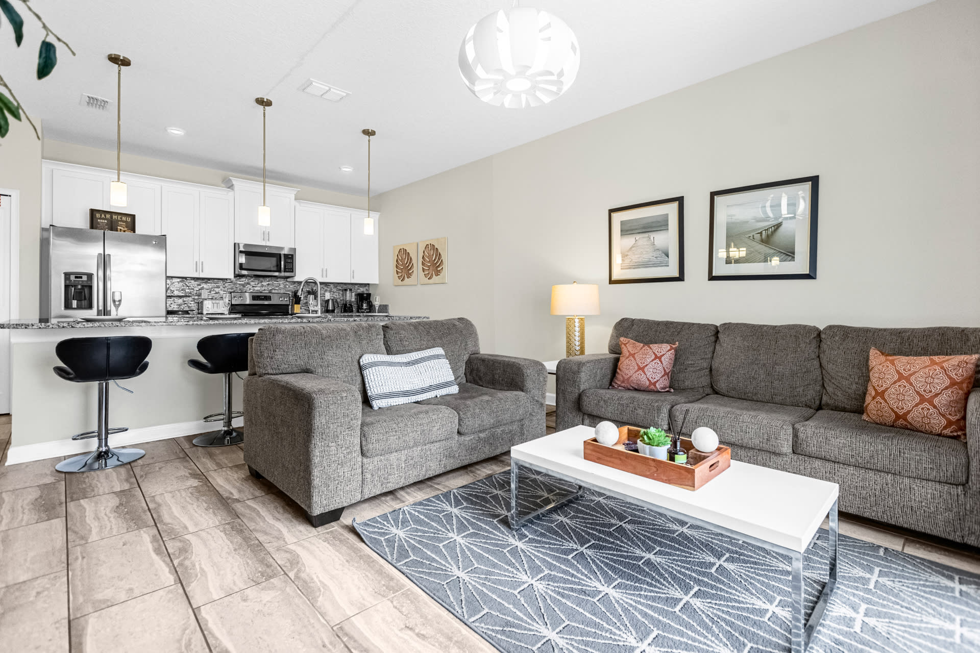 Open-concept living area featuring a gray sofa and loveseat, a white coffee table on a geometric rug, and a view into the modern kitchen with white cabinets and a breakfast bar.