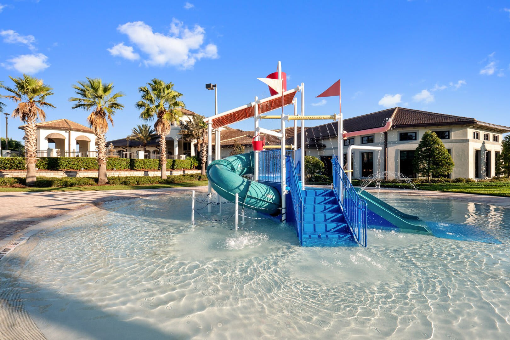 Shallow children's splash pad area with a blue and green water play structure, including a slide and water features. The area is surrounded by paved ground and resort buildings with palm trees in the background, perfect for young guests.