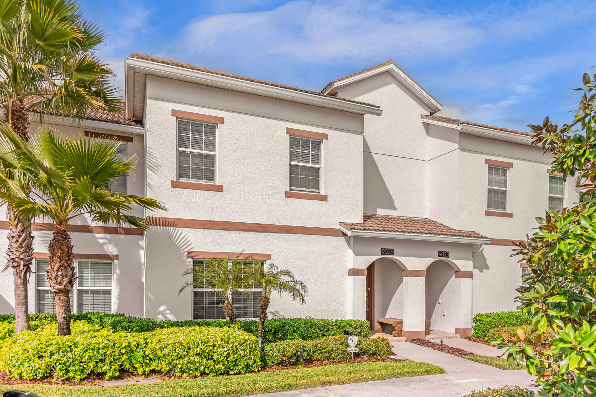 Front exterior of a two-story townhouse with light stucco walls and brown trim, featuring multiple windows with white blinds. Lush green landscaping and palm trees line the front entrance.