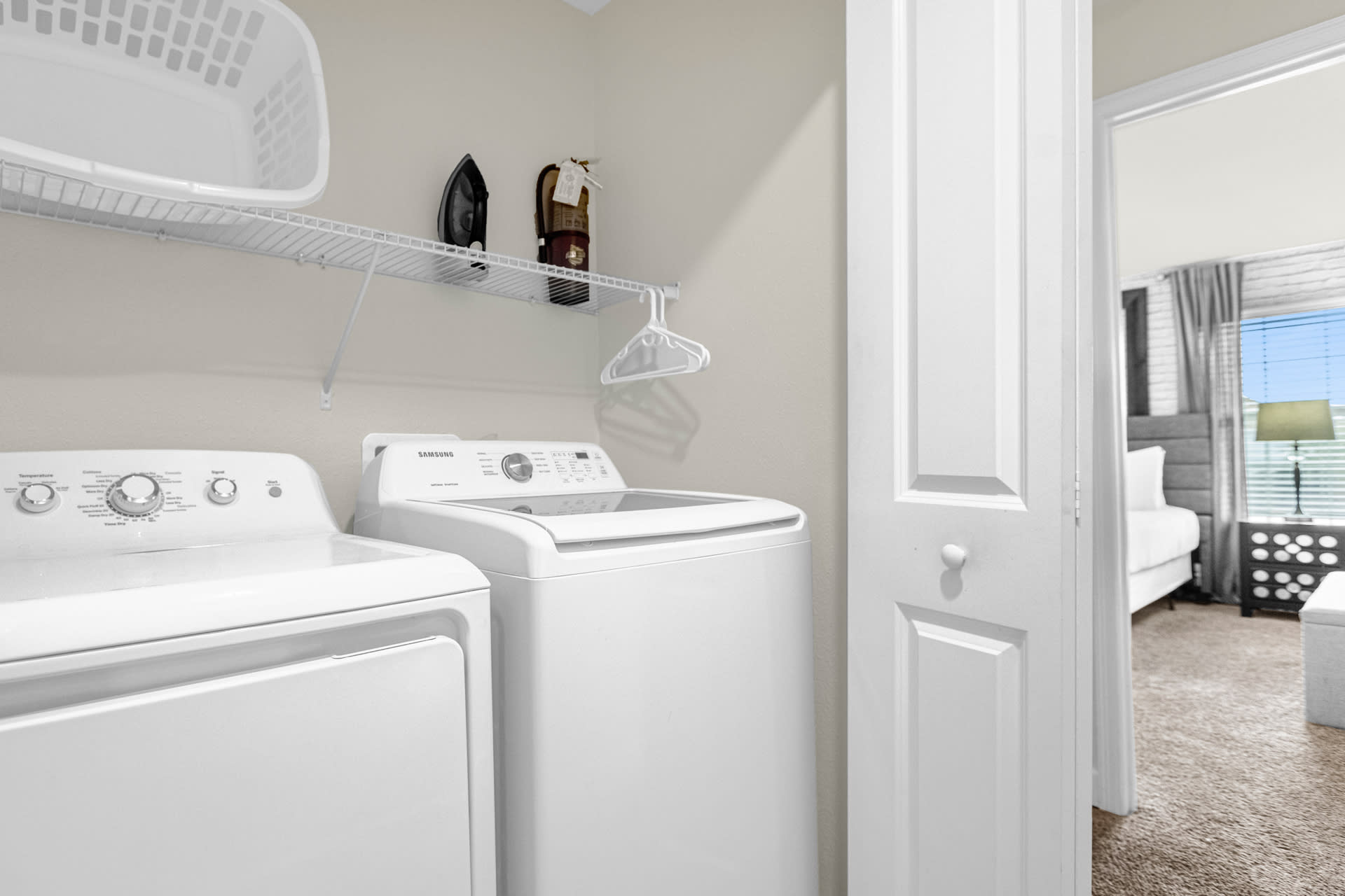 Compact laundry closet with a white top-loading washing machine and a front-loading dryer. A wire shelf above holds a laundry basket, iron, and fire extinguisher, with a white hanger visible next to an open door leading to another room.