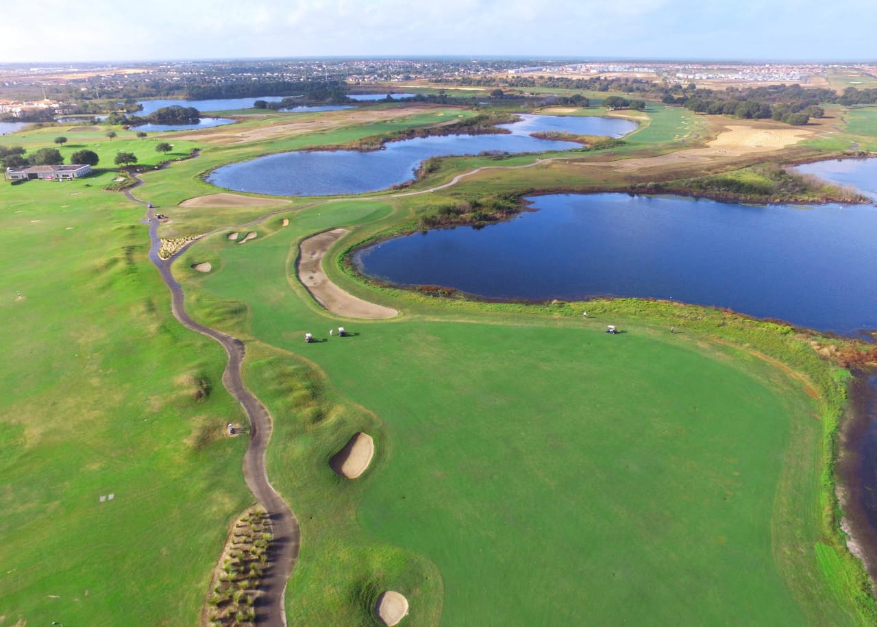 An expansive aerial view of a lush green golf course featuring multiple water hazards and sand traps, winding through a developed landscape under a partly cloudy sky. Golf carts are visible on the course.