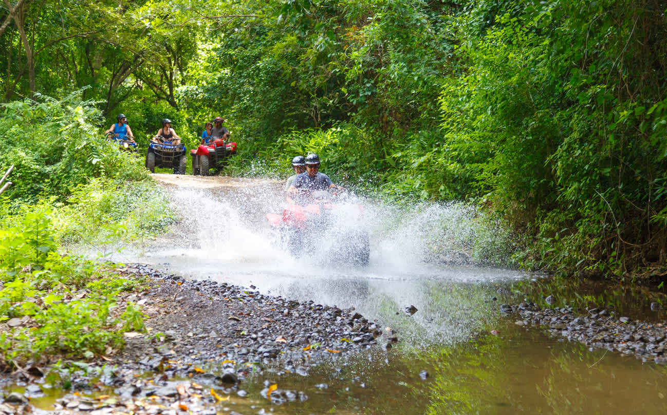 Several people on ATVs ride through a shallow, rocky stream in a dense, vibrant green forest, creating large splashes of water. The scene suggests an adventurous outdoor activity.