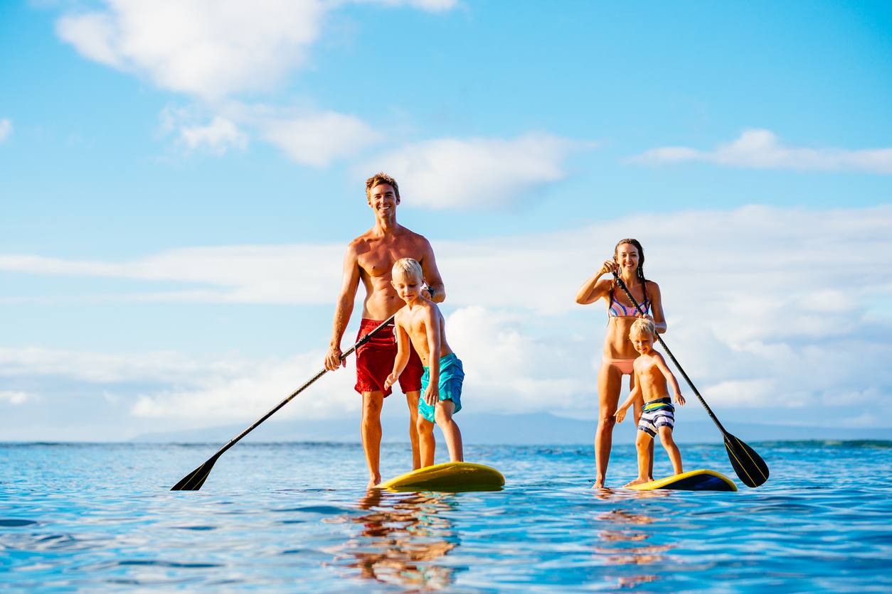 A family of four, including two adults and two young children, stands on paddleboards in calm, clear blue ocean water under a bright, partly cloudy sky. They are smiling and holding paddles.
