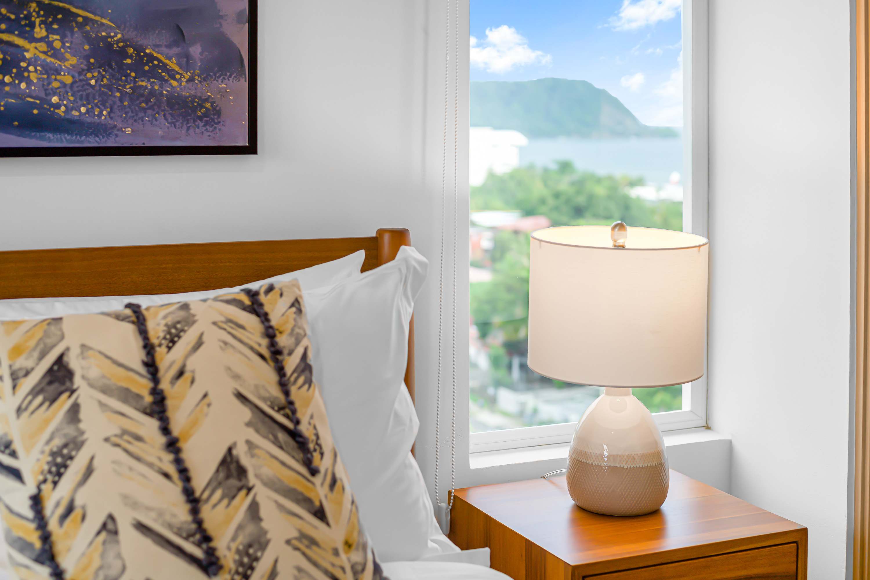Close-up of a bedside table with a modern lamp, next to a bed with white linens and a decorative pillow, with a window revealing a partial sea view and lush greenery outside.