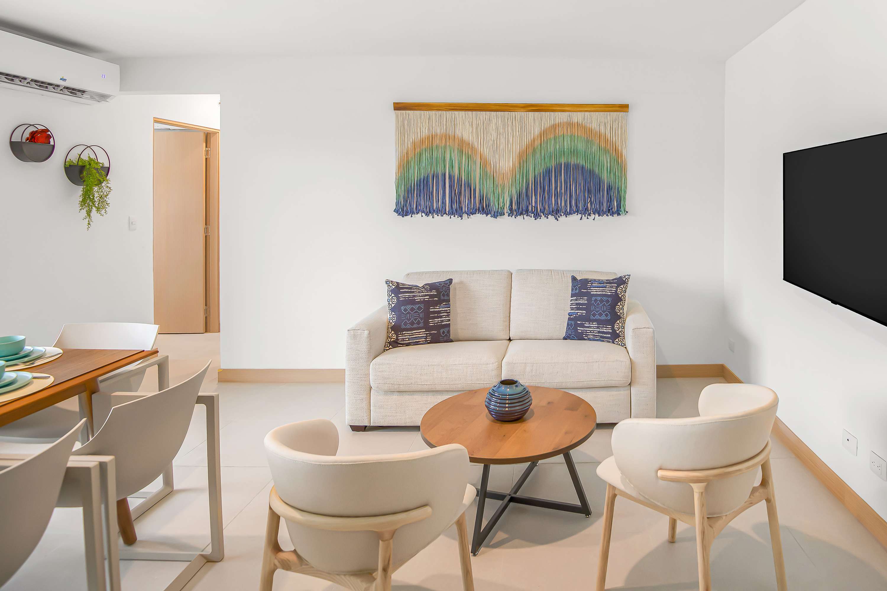 Modern living room with a cream sofa, two light-colored accent chairs, and a round wooden coffee table with a decorative vase. A large colorful tapestry hangs above the sofa, and a wall-mounted television is visible on the right.