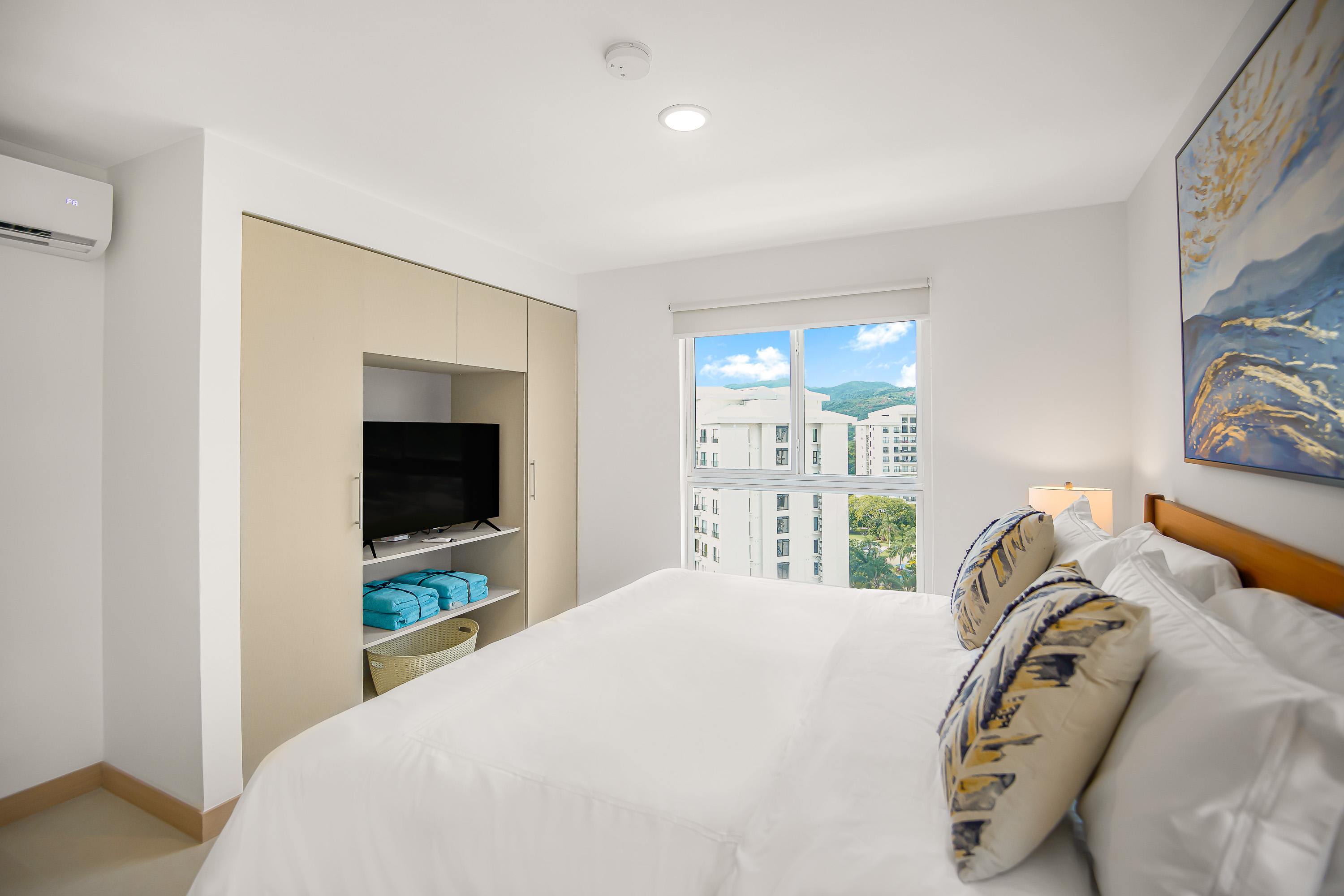 Bright bedroom featuring a large bed with white linens and decorative pillows, a built-in light wood wardrobe with a television nook, and a large window offering views of buildings and distant mountains.