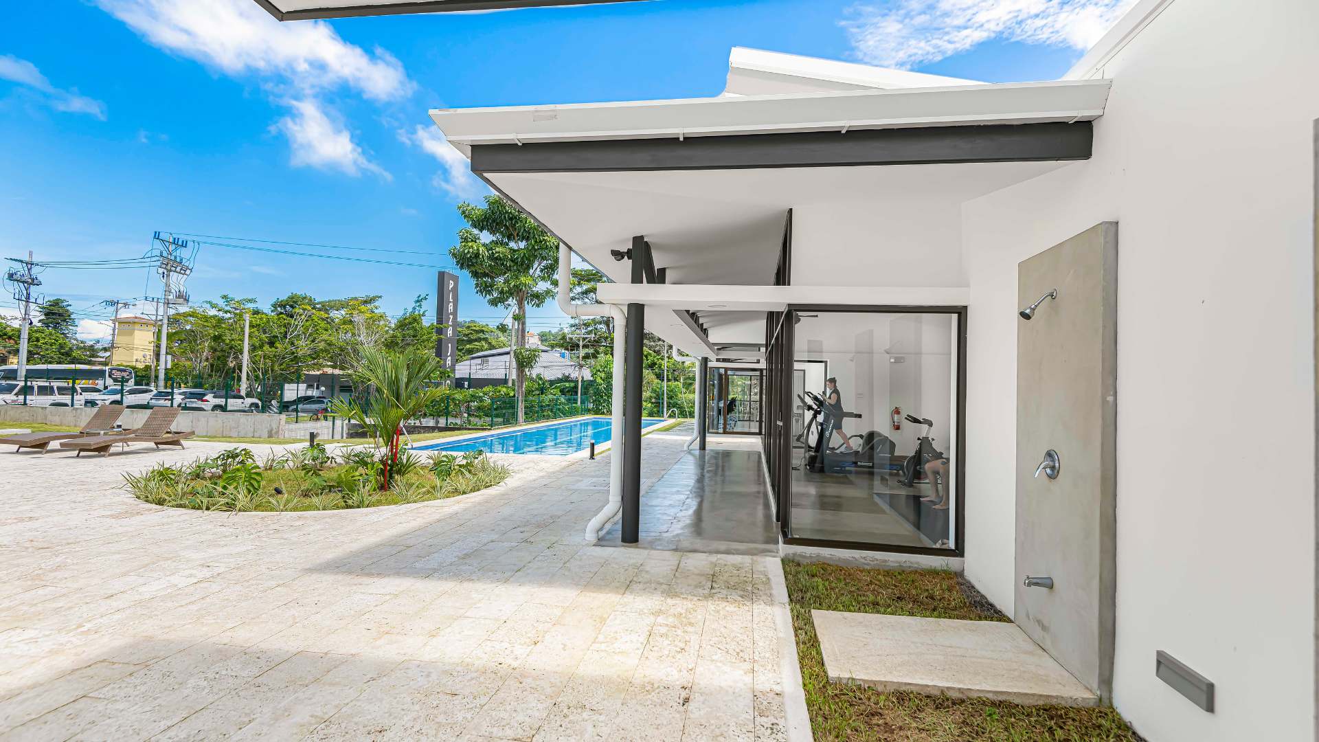Outdoor view of a modern gym facility with large glass windows, showing exercise equipment inside, adjacent to a swimming pool area with lounge chairs and lush landscaping.