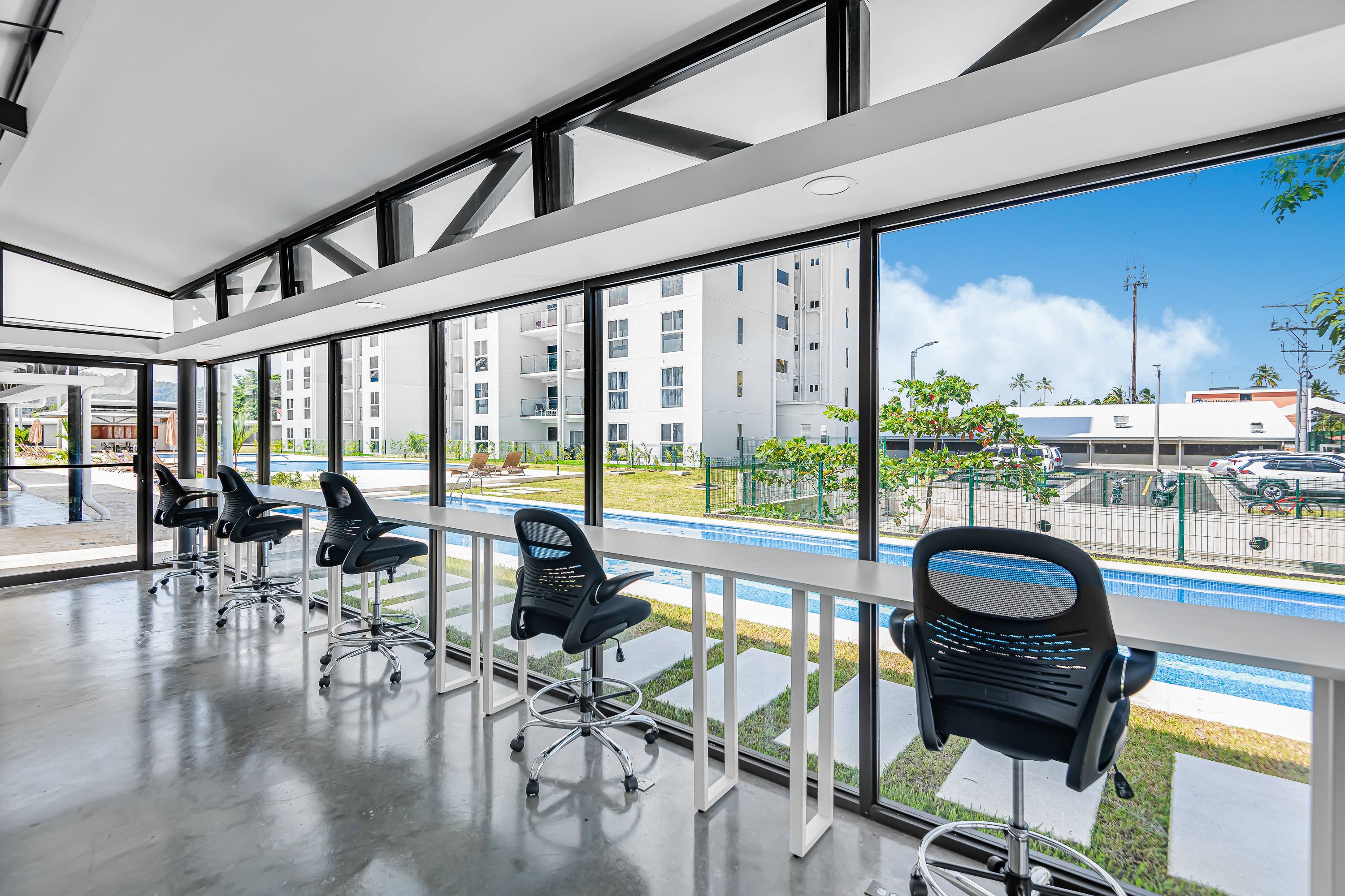 Modern co-working space with a long white desk and multiple black swivel chairs, offering panoramic views of a swimming pool and surrounding buildings through large windows. The floor is polished concrete.