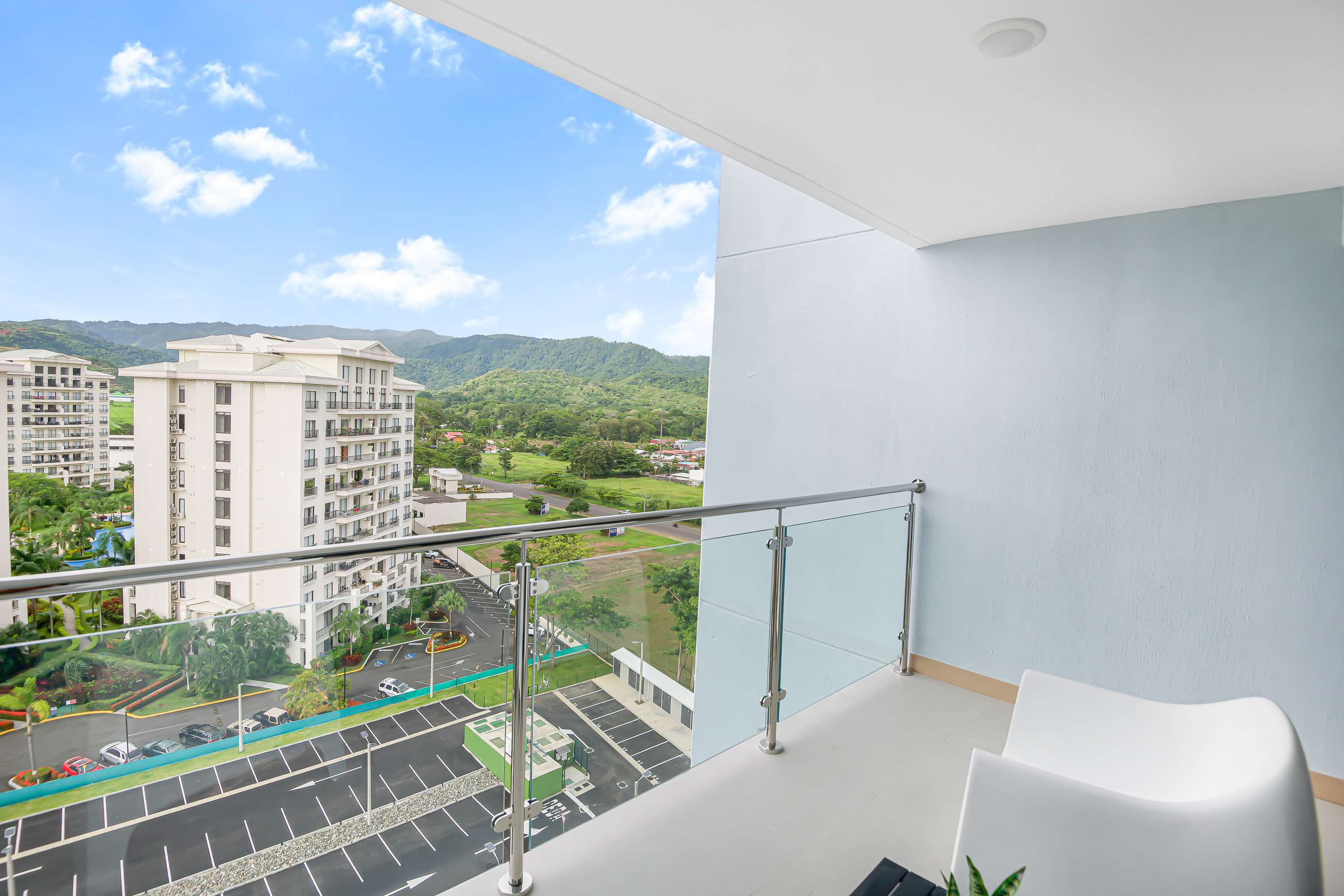 Balcony with glass railings and light blue walls, offering a wide view of the complex's parking lot, other buildings, and distant green mountains under a bright blue sky. A small plant sits on a black table next to a white lounge chair.