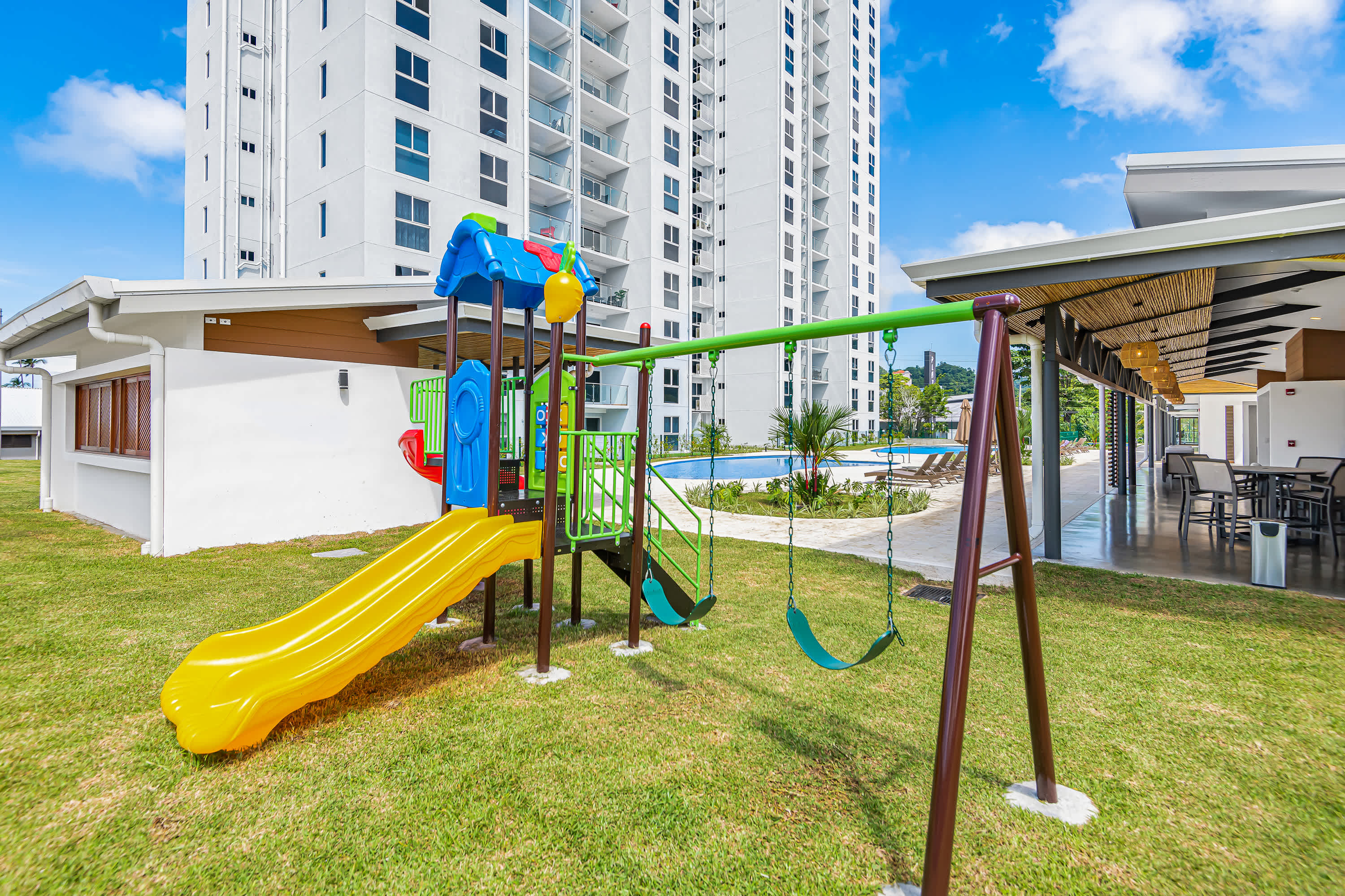 Vibrant outdoor playground featuring a colorful slide and swings on a grassy lawn, with a swimming pool and tall residential buildings in the background. The area is family-friendly and well-maintained.