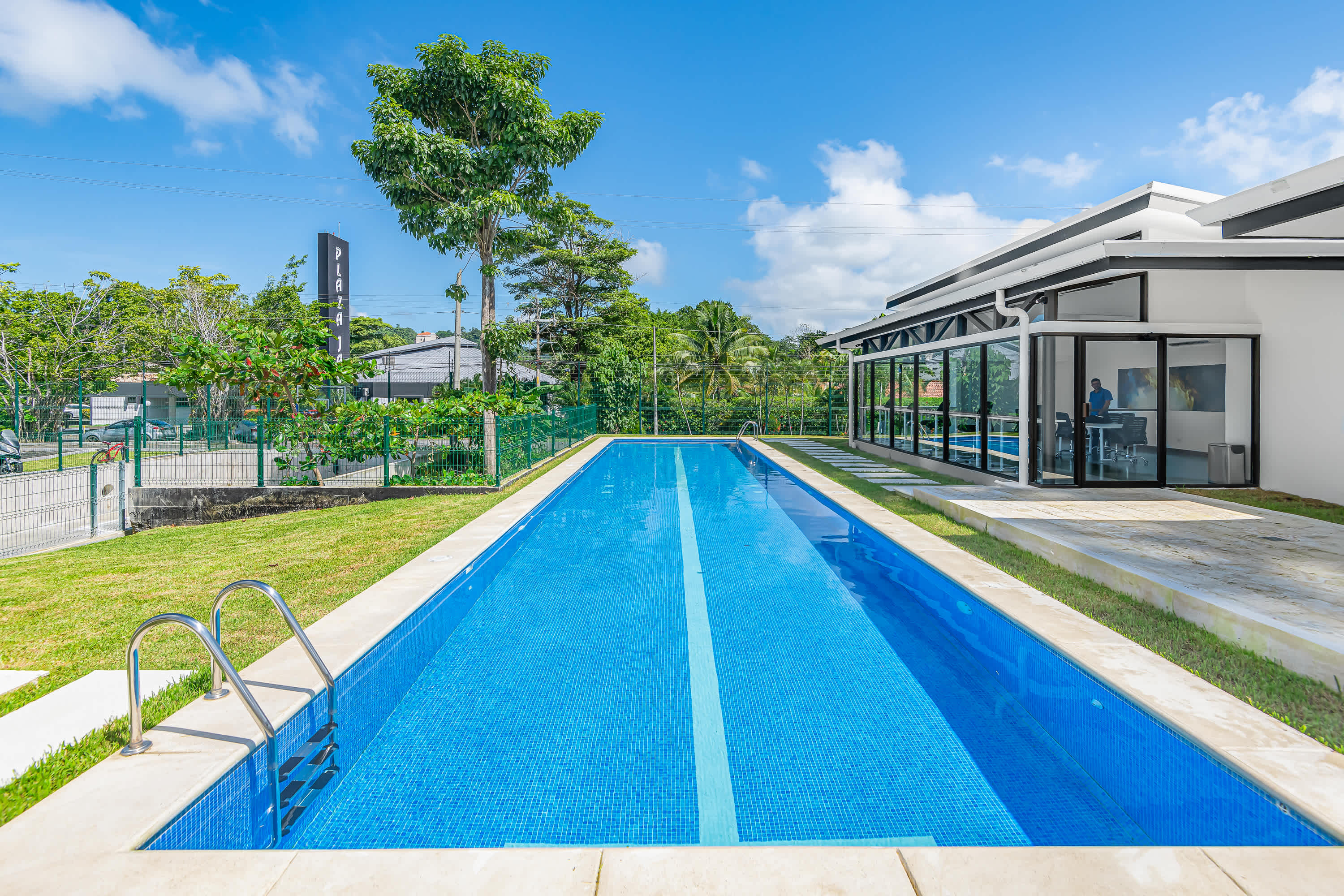 A long, narrow outdoor swimming pool with vibrant blue mosaic tiles and a white lane line is set in a manicured green lawn. A modern building with large windows and a white exterior stands beside it under a clear blue sky.