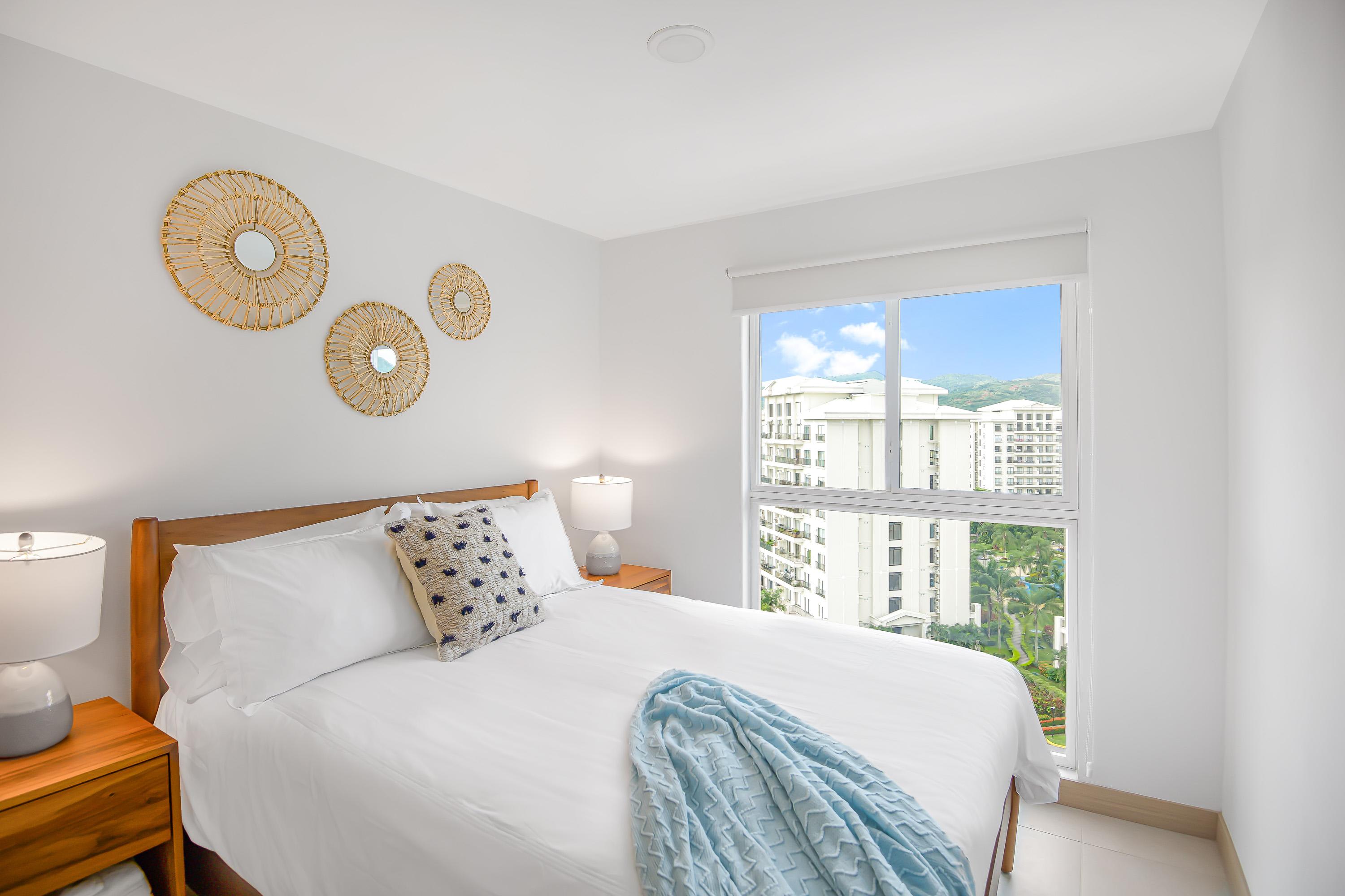 Bright bedroom featuring a queen-sized bed with white linens, two wooden nightstands with lamps, and decorative woven wall art. A large window offers views of modern buildings and distant green hills.