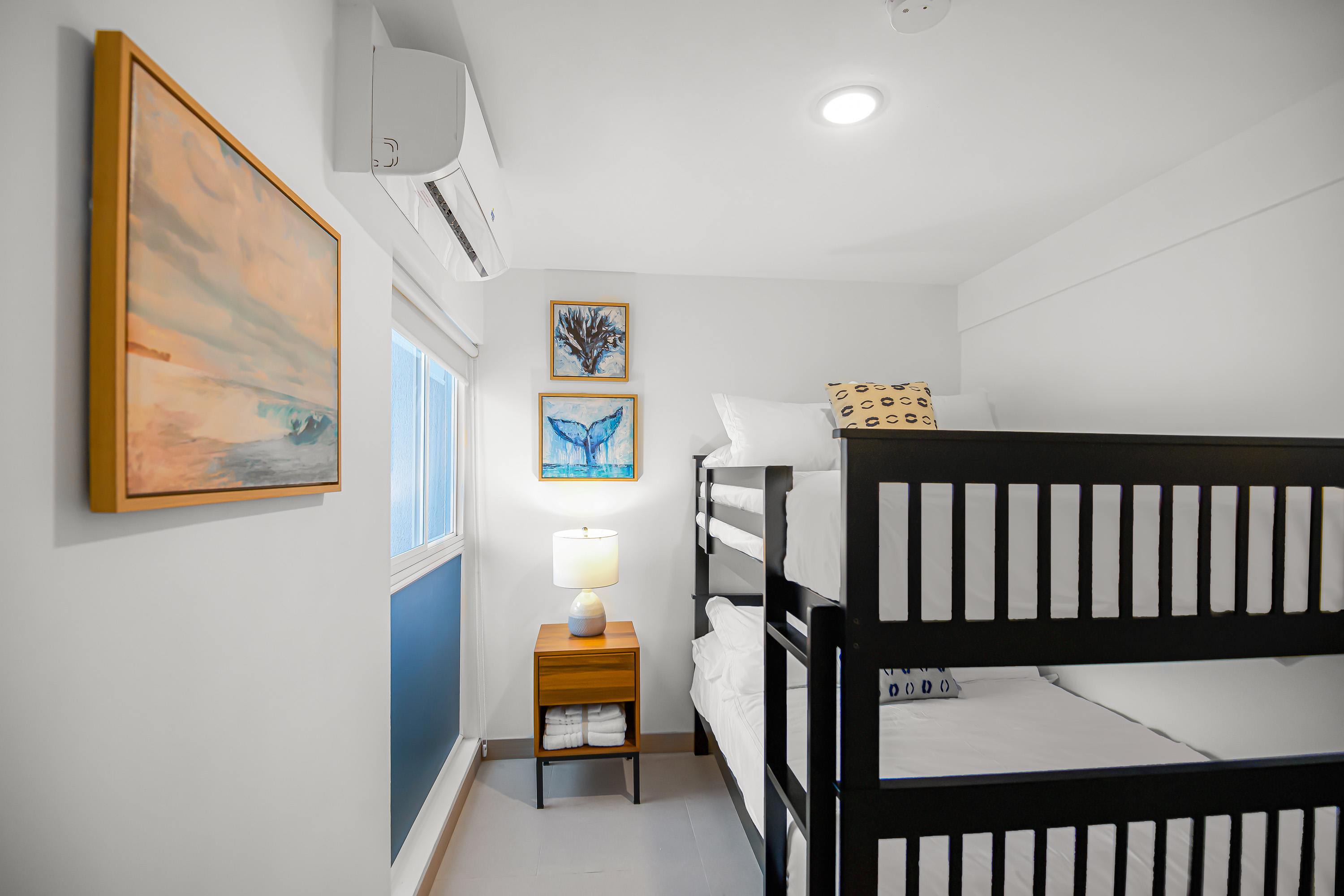 Compact bedroom featuring black wooden bunk beds with white bedding, a small wooden nightstand with a lamp, and two framed artworks on the wall. A window with room-darkening shades is visible.