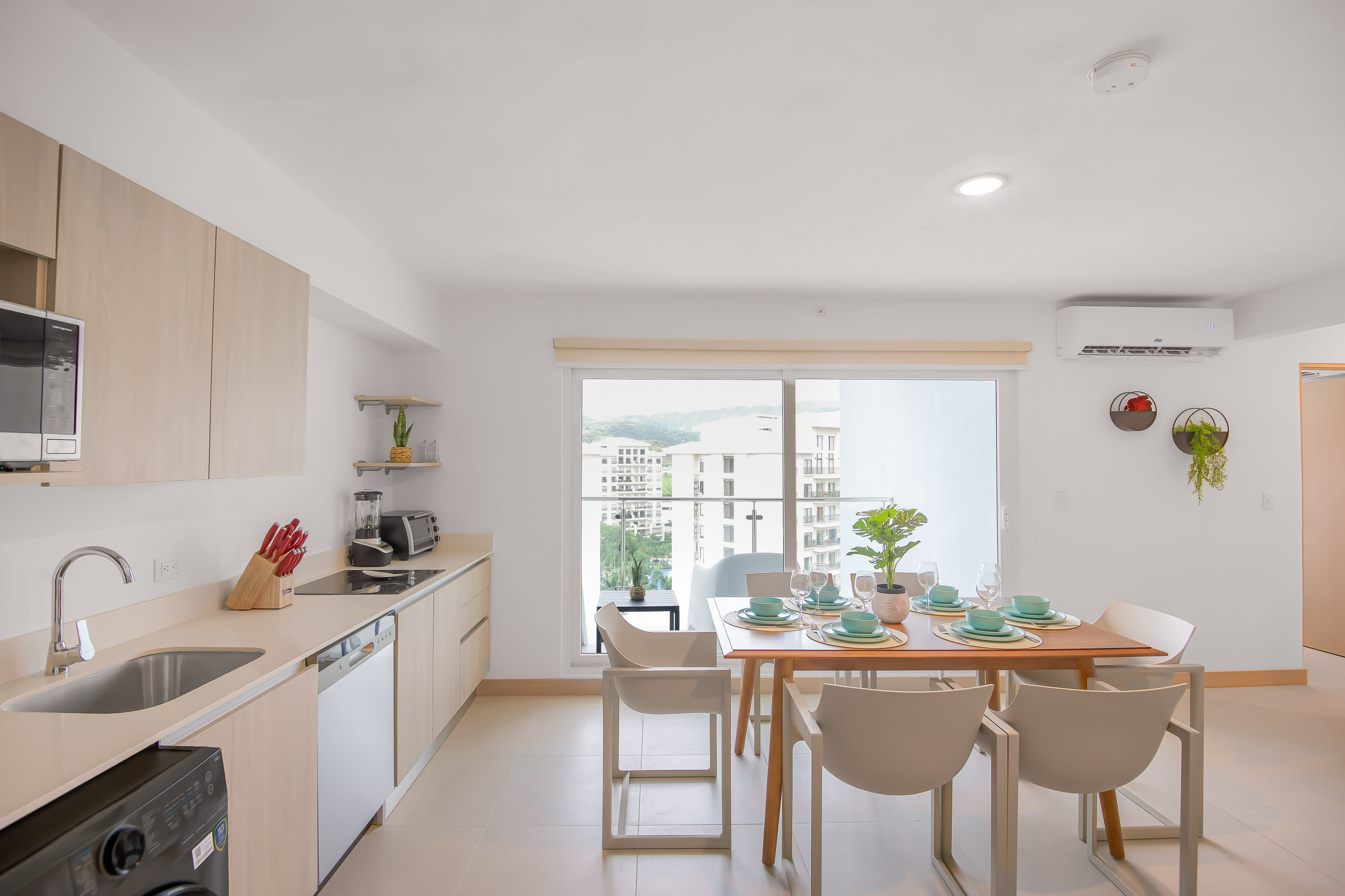 Open-concept dining area with a light wood table and six white chairs, set with blue and white dishes, adjacent to a kitchen with light wood cabinets and a sliding glass door leading to a balcony with city views.