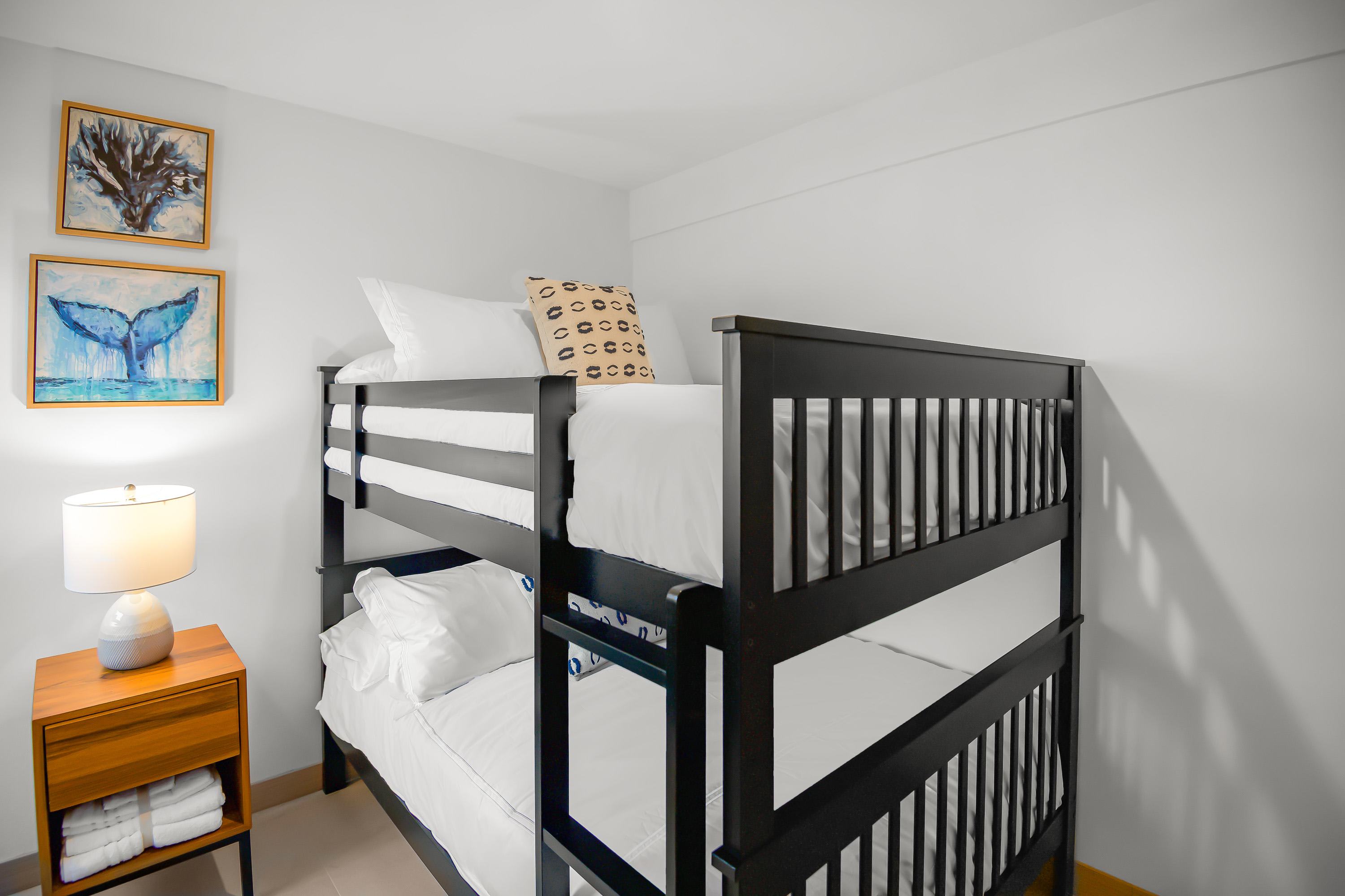 Close-up view of the black wooden bunk beds with white linens and decorative pillows. Two framed artworks depicting marine life hang above a small wooden nightstand with a lamp.