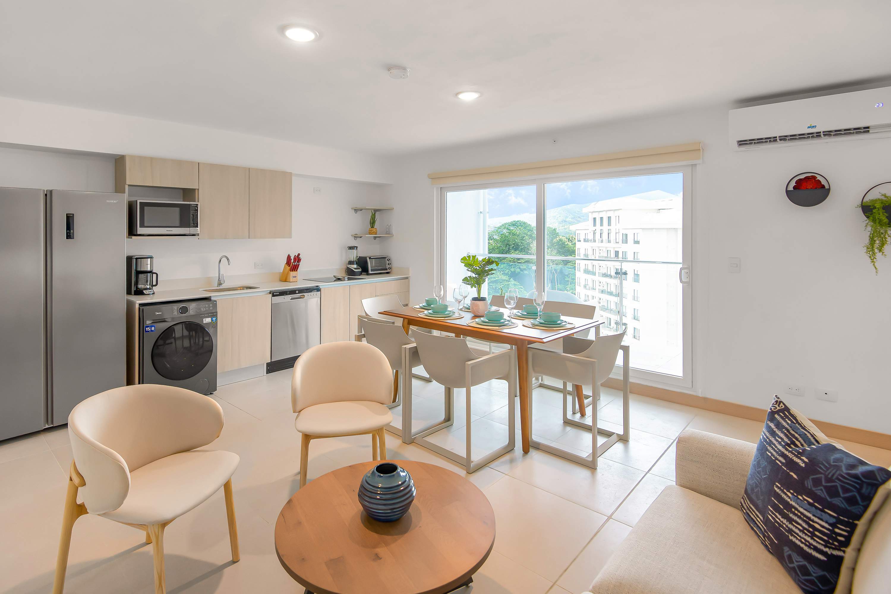 Open-concept living, dining, and kitchen area featuring a cream sofa, two accent chairs, a round wooden coffee table, and a dining table set for six. The modern kitchen has light wood cabinets and stainless steel appliances, with a large sliding glass door leading to a balcony.
