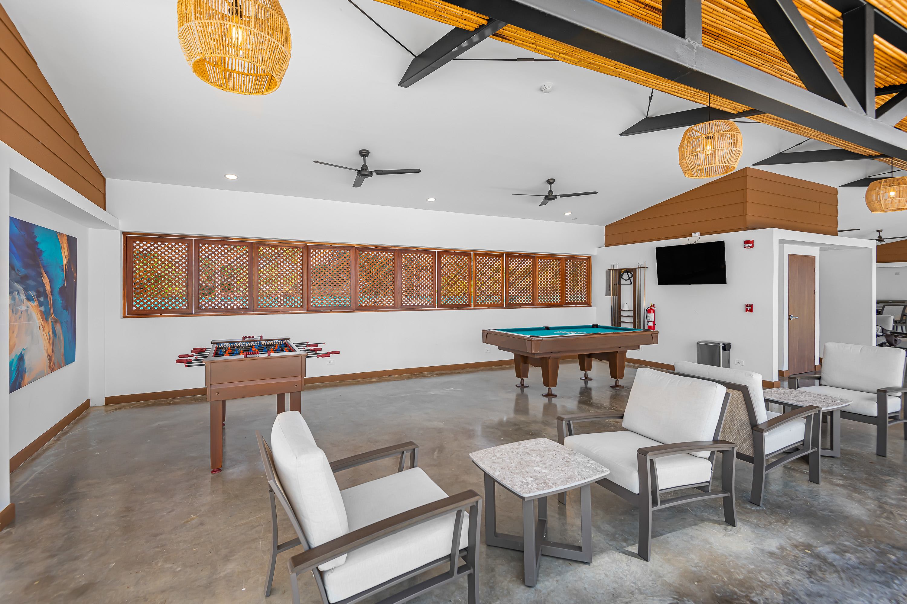 Spacious recreation room with a foosball table, a pool table, comfortable armchairs with small tables, and decorative woven light fixtures, featuring a high ceiling with exposed beams.
