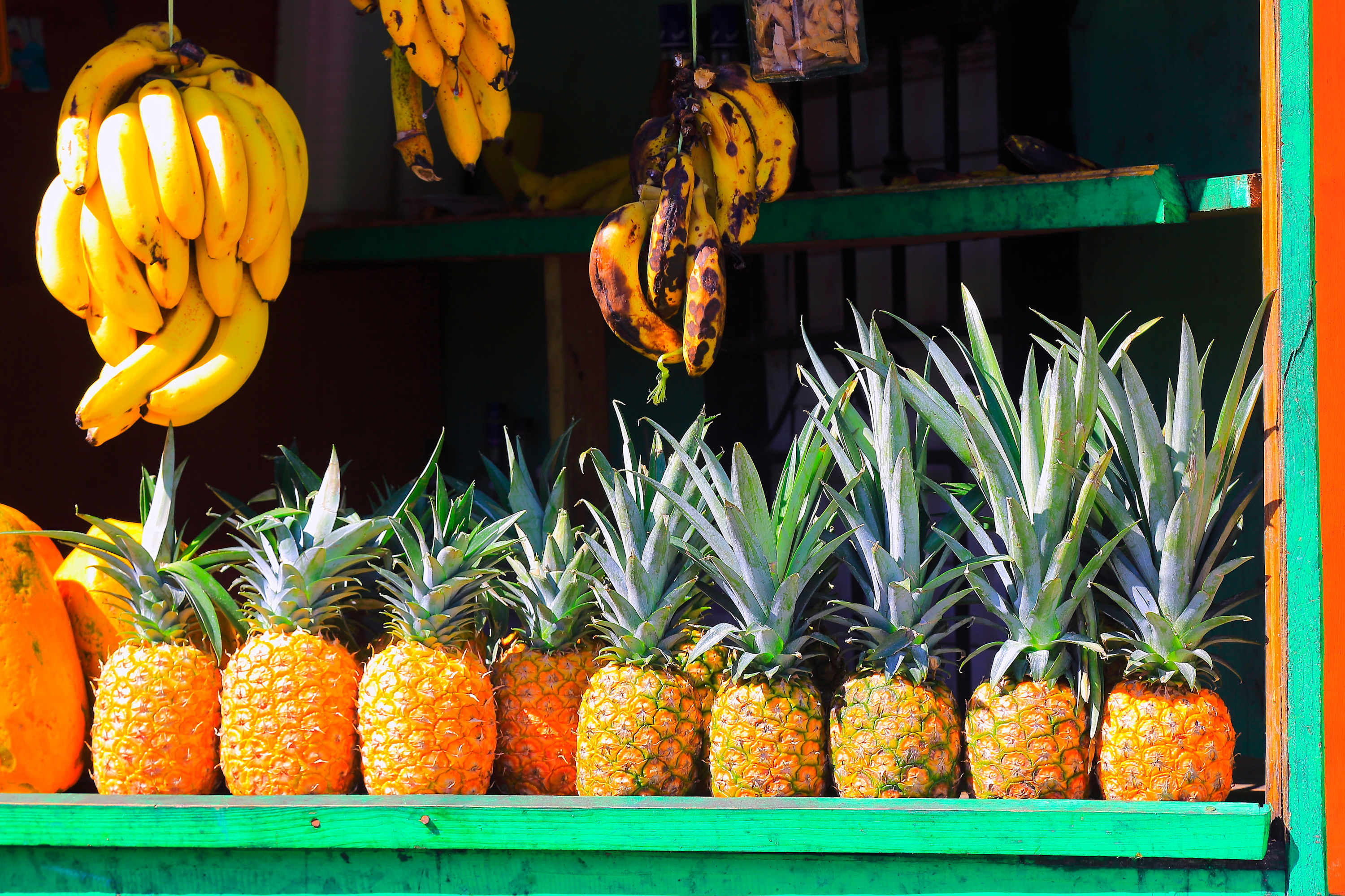 A vibrant market stall displays rows of fresh, ripe pineapples on a green shelf, with large bunches of yellow bananas hanging above. The scene suggests local produce and a tropical setting.