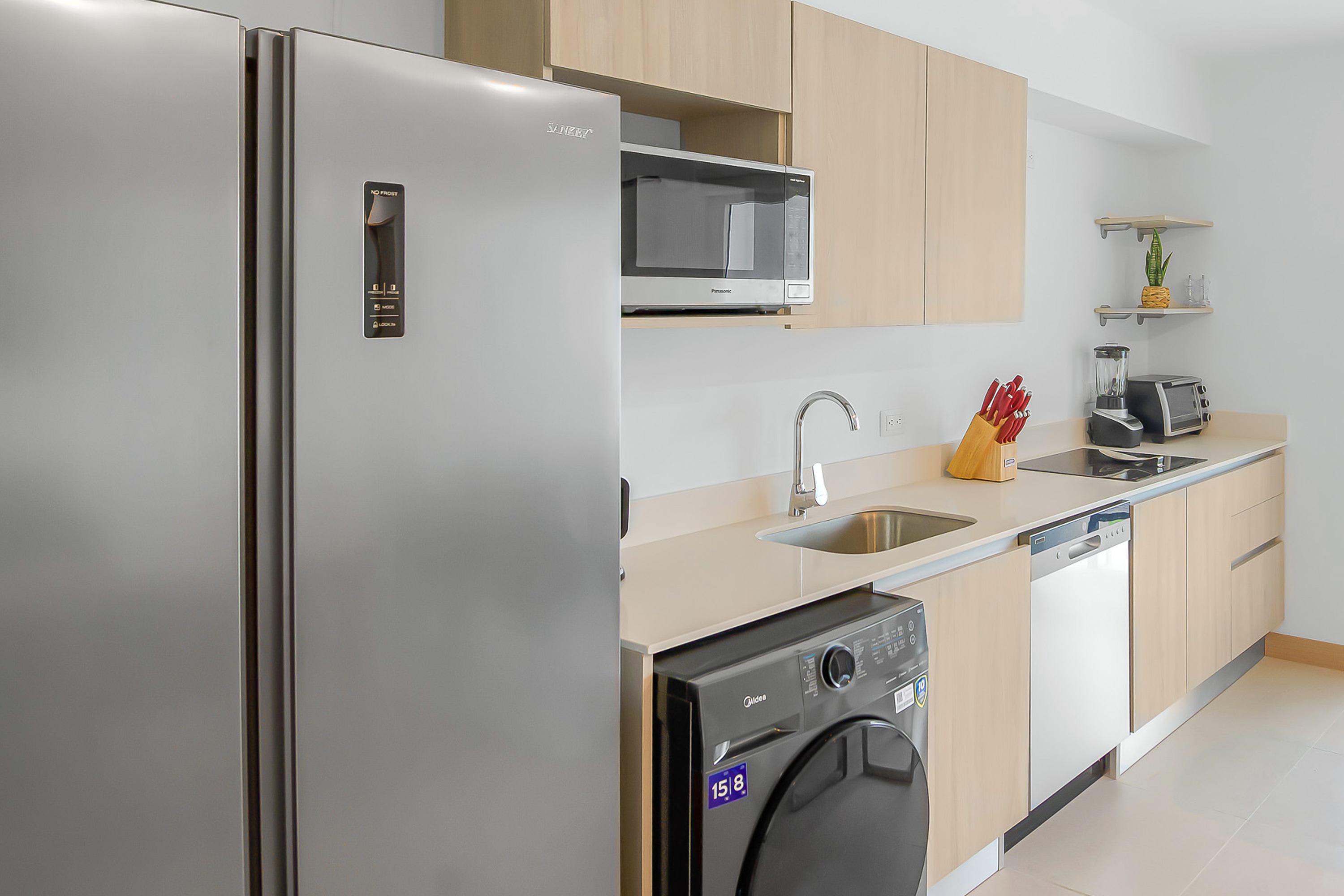 Modern kitchen with light wood cabinetry, stainless steel appliances including a refrigerator, microwave, and dishwasher, a sink, and a black front-load washing machine integrated below the counter.