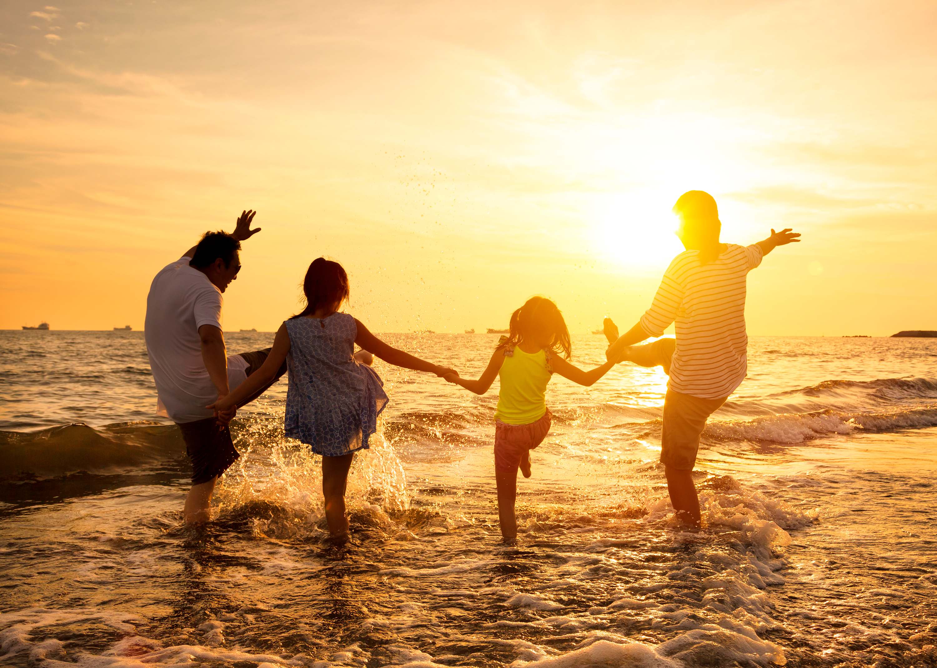 A family of four, silhouetted against a golden sunset, holds hands and splashes playfully in the shallow ocean waves on a sandy beach. The warm light reflects on the water.