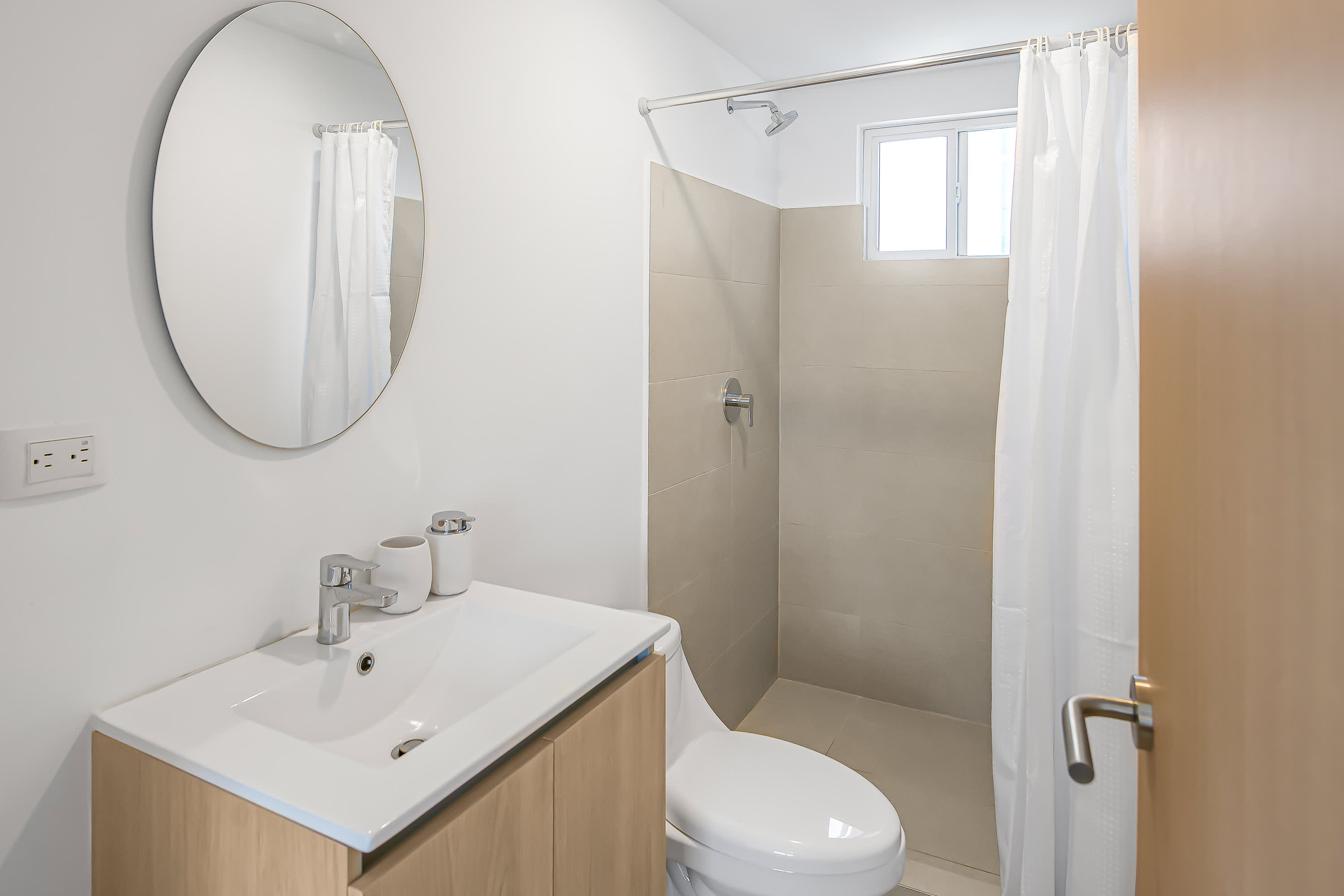 Modern bathroom with a light wood vanity, white rectangular sink, and a round mirror above. The shower features light beige tiled walls and a white shower curtain, with a toilet beside it.