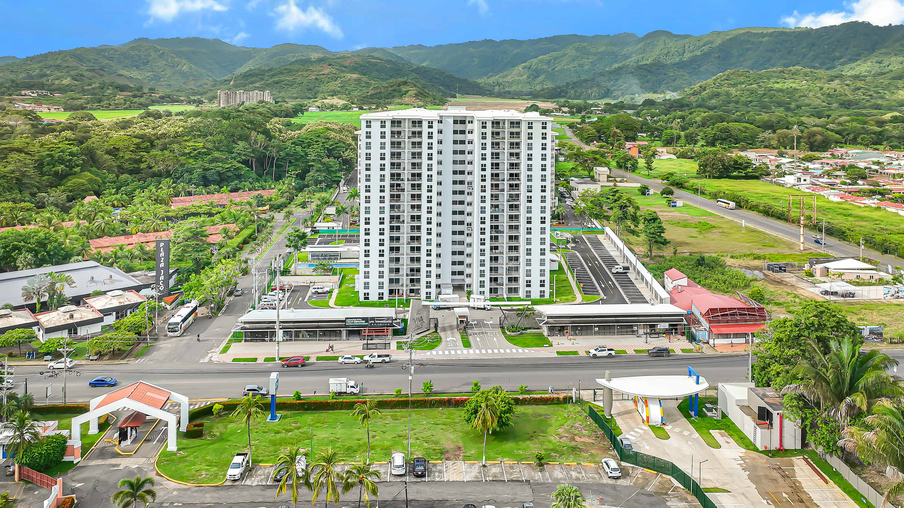 Aerial view of a tall, modern white condominium building surrounded by lush green mountains and a mix of residential and commercial areas, with a road and parking lots visible.