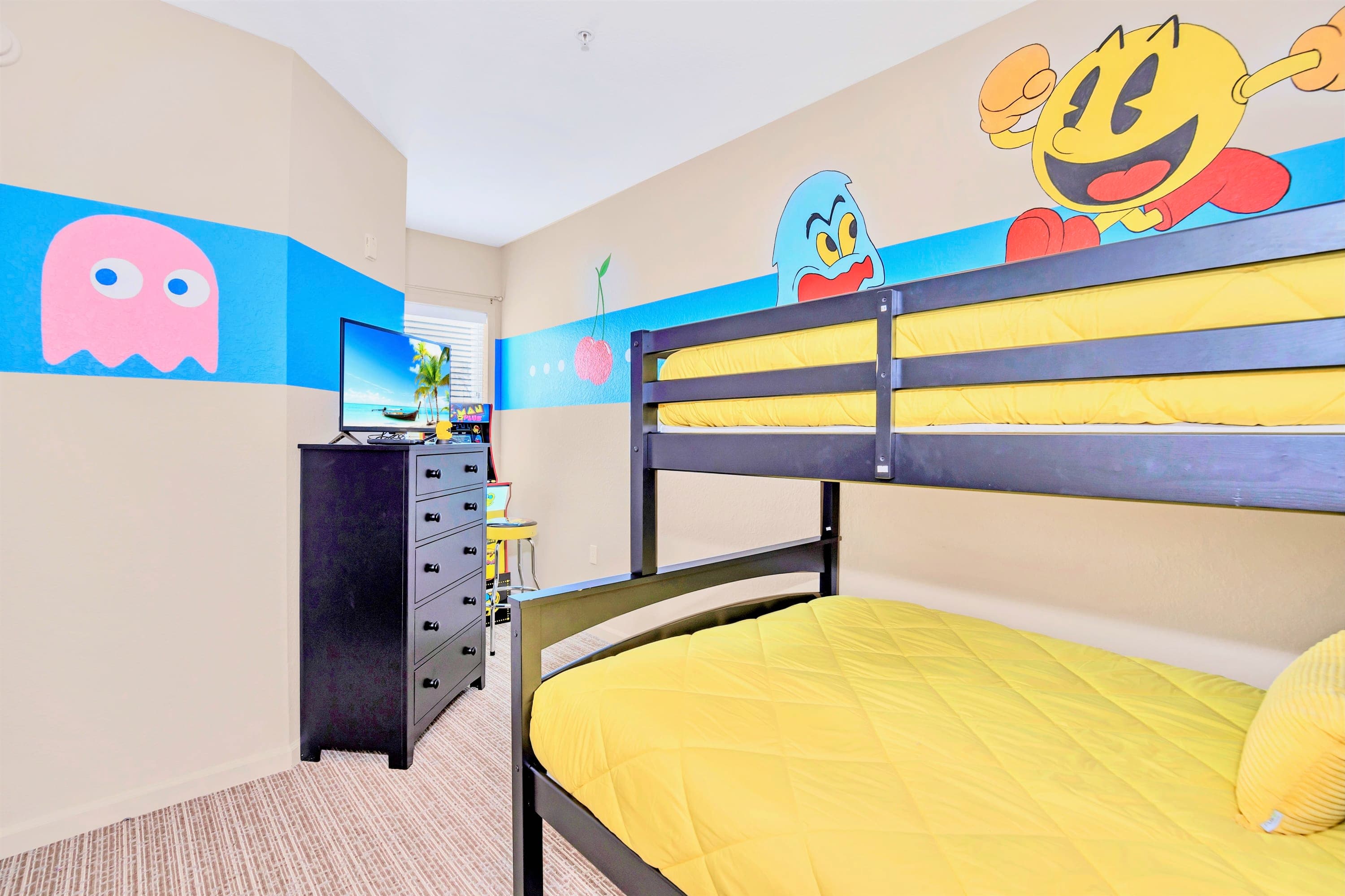 View of the Pac-Man themed children's bedroom, showing the black bunk bed with yellow bedding and a black dresser with a TV displaying a beach scene. The walls are decorated with a blue stripe and Pac-Man characters.