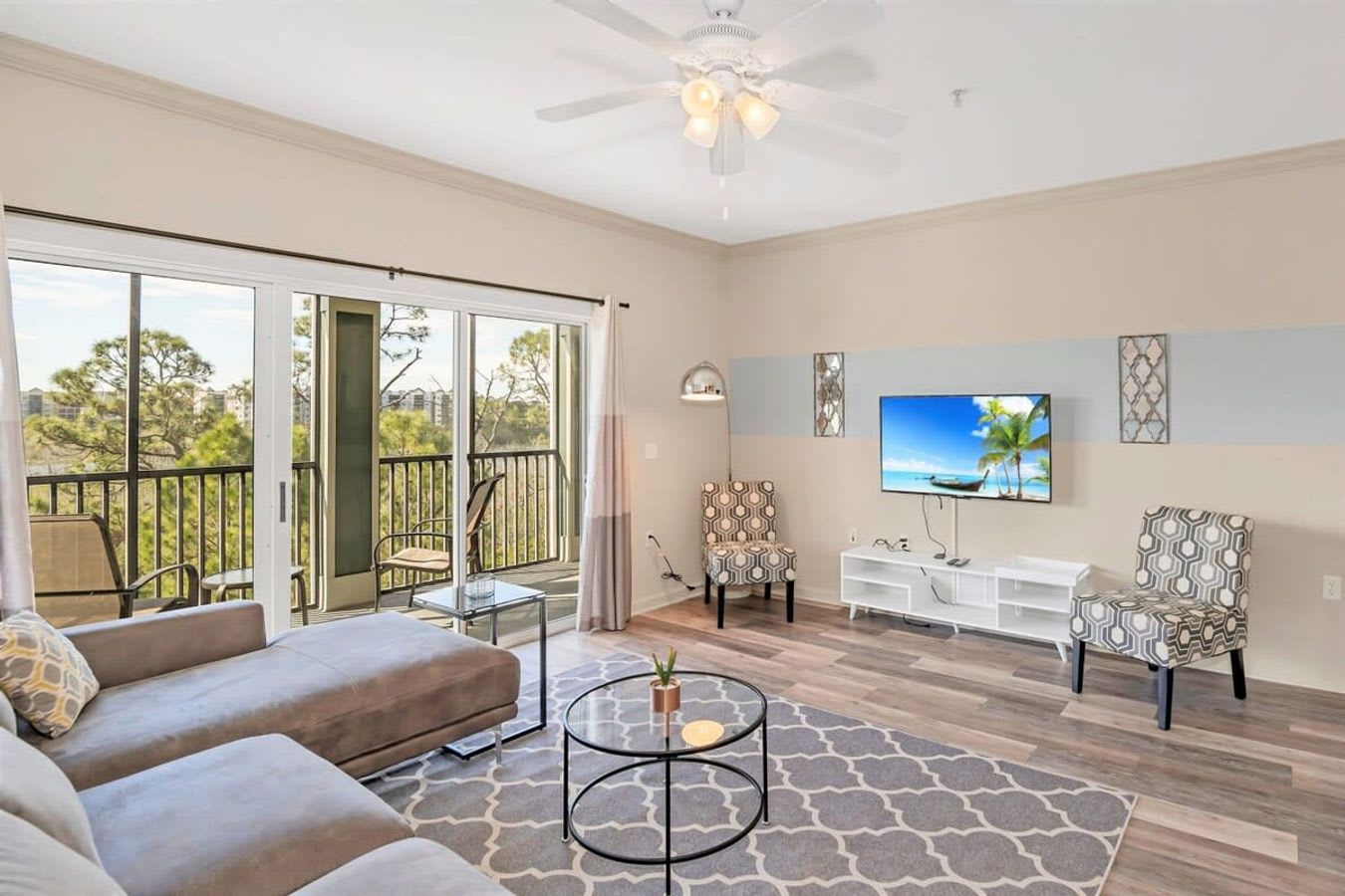 Bright living room with a large L-shaped sectional sofa, two round glass coffee tables, and a patterned area rug on light wood-look flooring. A wall-mounted TV is visible, and sliding glass doors lead to a balcony with outdoor seating.